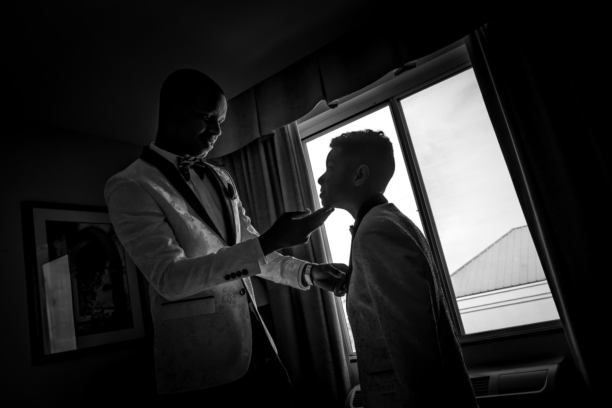 Black and white photograph of one groom helping the other with his jacket beside a bright hotel window
