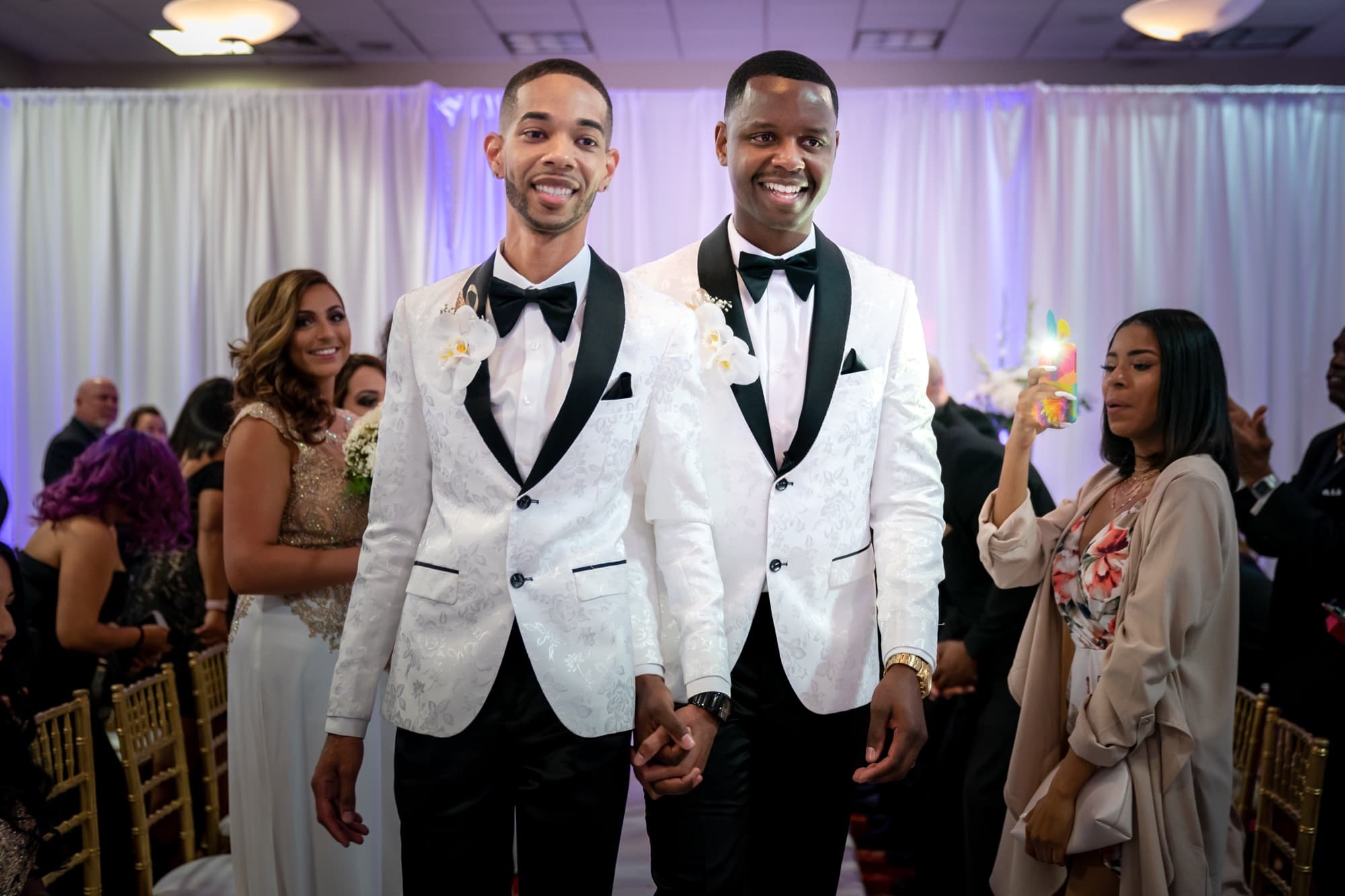 The grooms walking hand in hand up the aisle in matching white shawl-collar tuxedos as guests smile around them