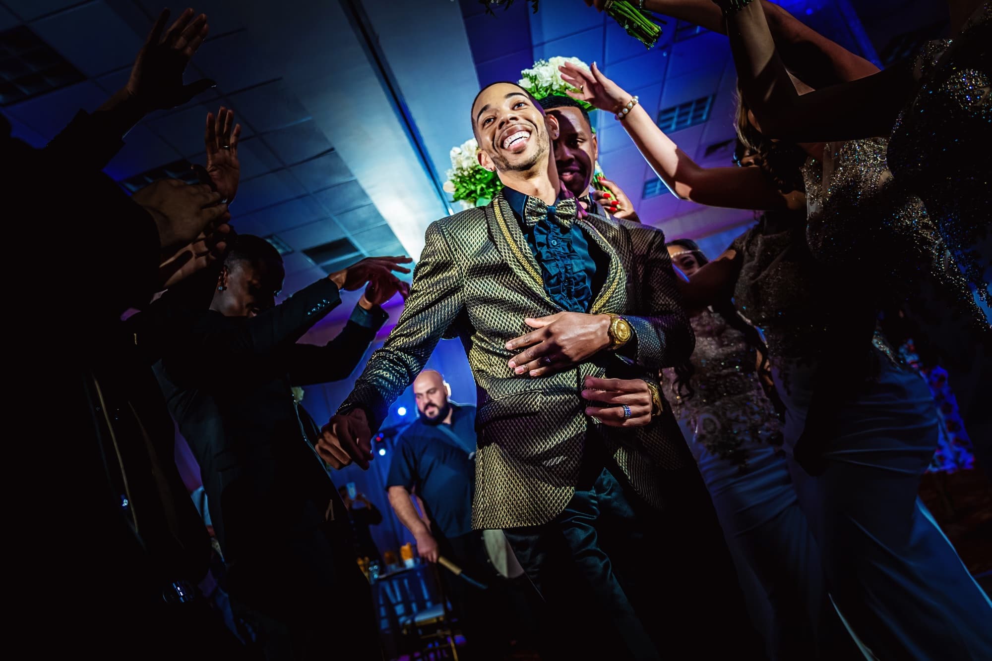 A groom in a textured gold and black jacket dancing in the middle of the reception crowd