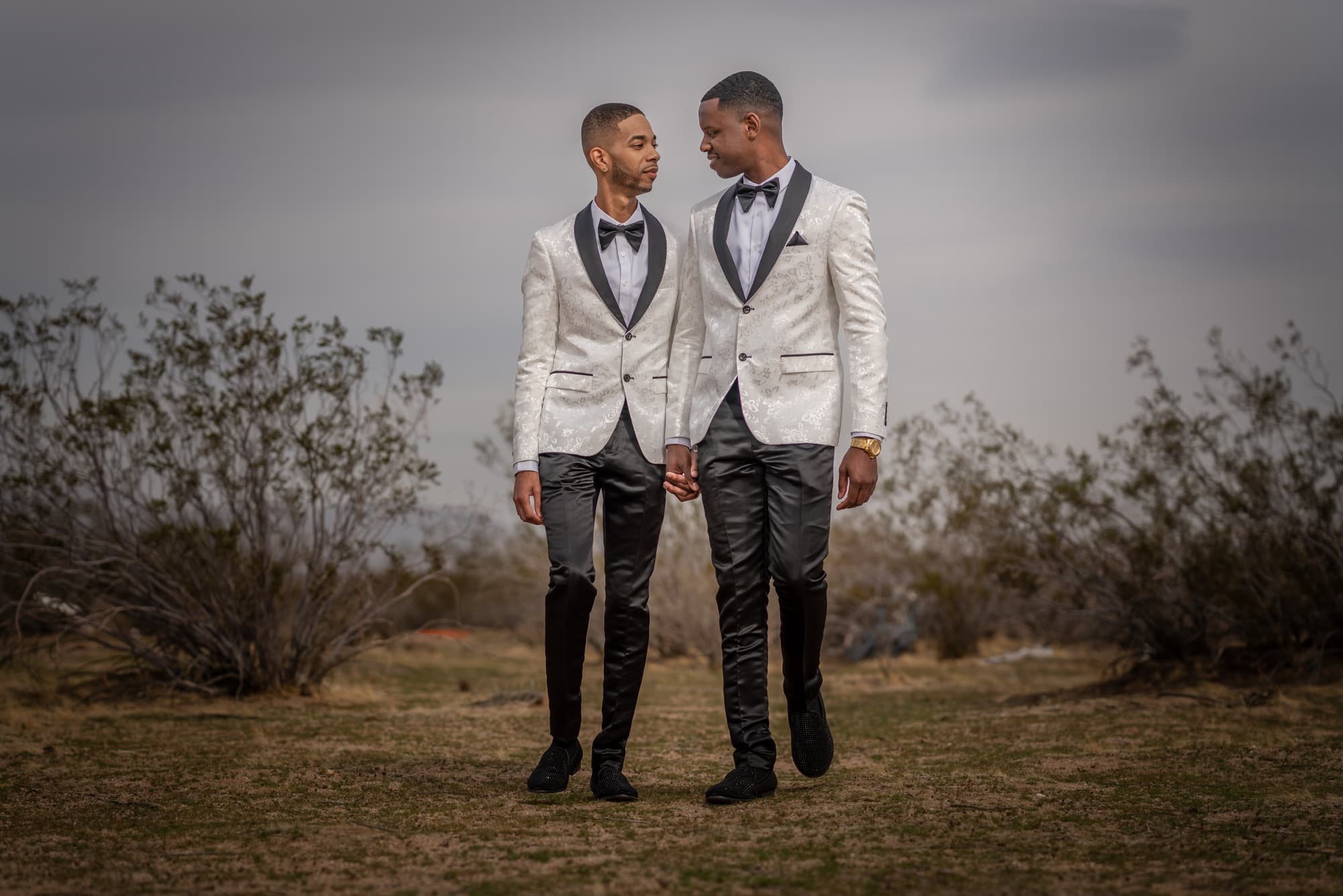 Anthony and Desmond walking hand in hand through High Desert scrub near Victorville in matching white shawl-collar tuxedos under a moody gray sky, photographed by Michael Anthony Photography