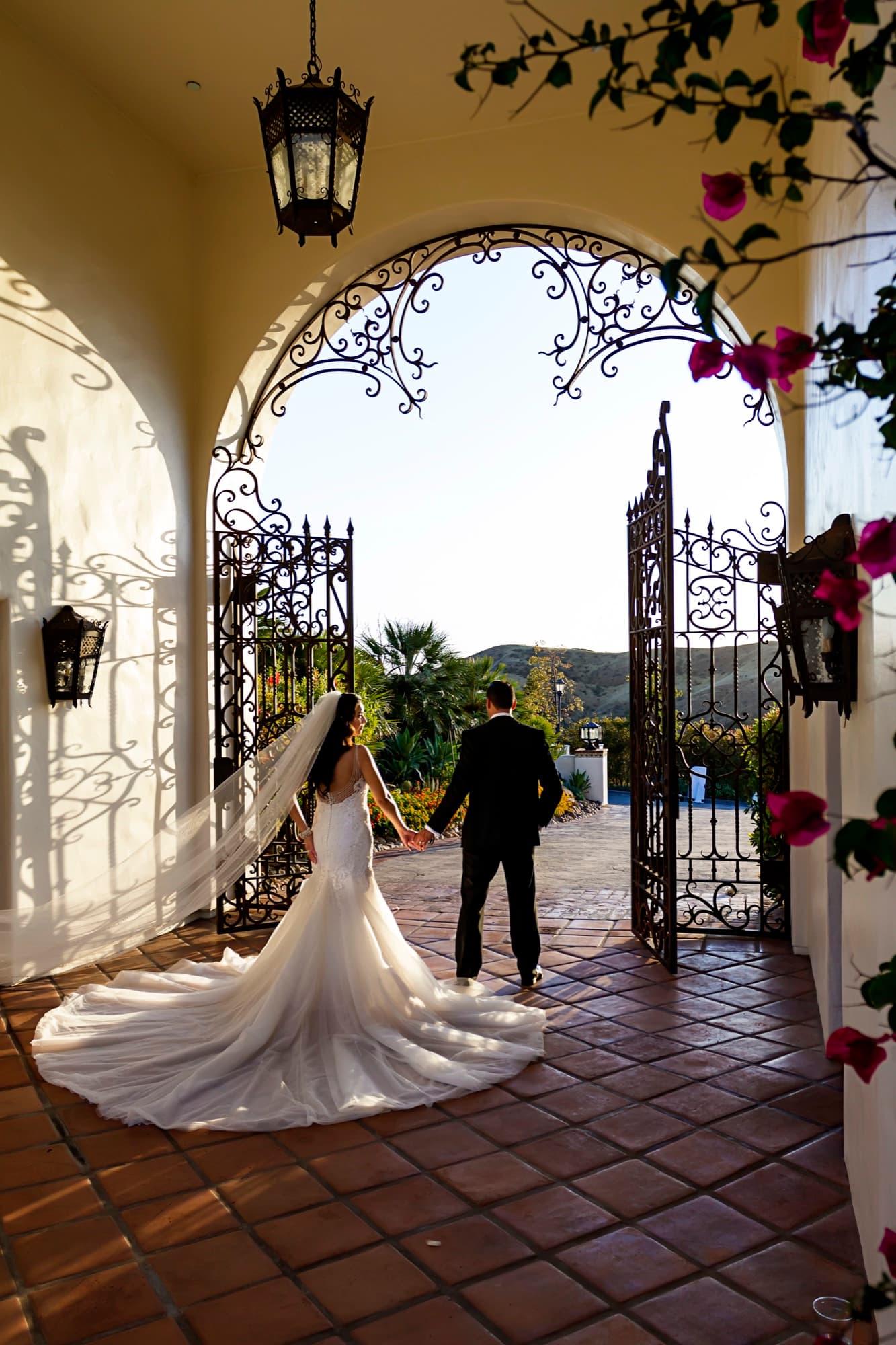 Bride's lace train spread across the terracotta tile as the couple walks hand in hand through the bougainvillea-framed gate at Hummingbird Nest Ranch