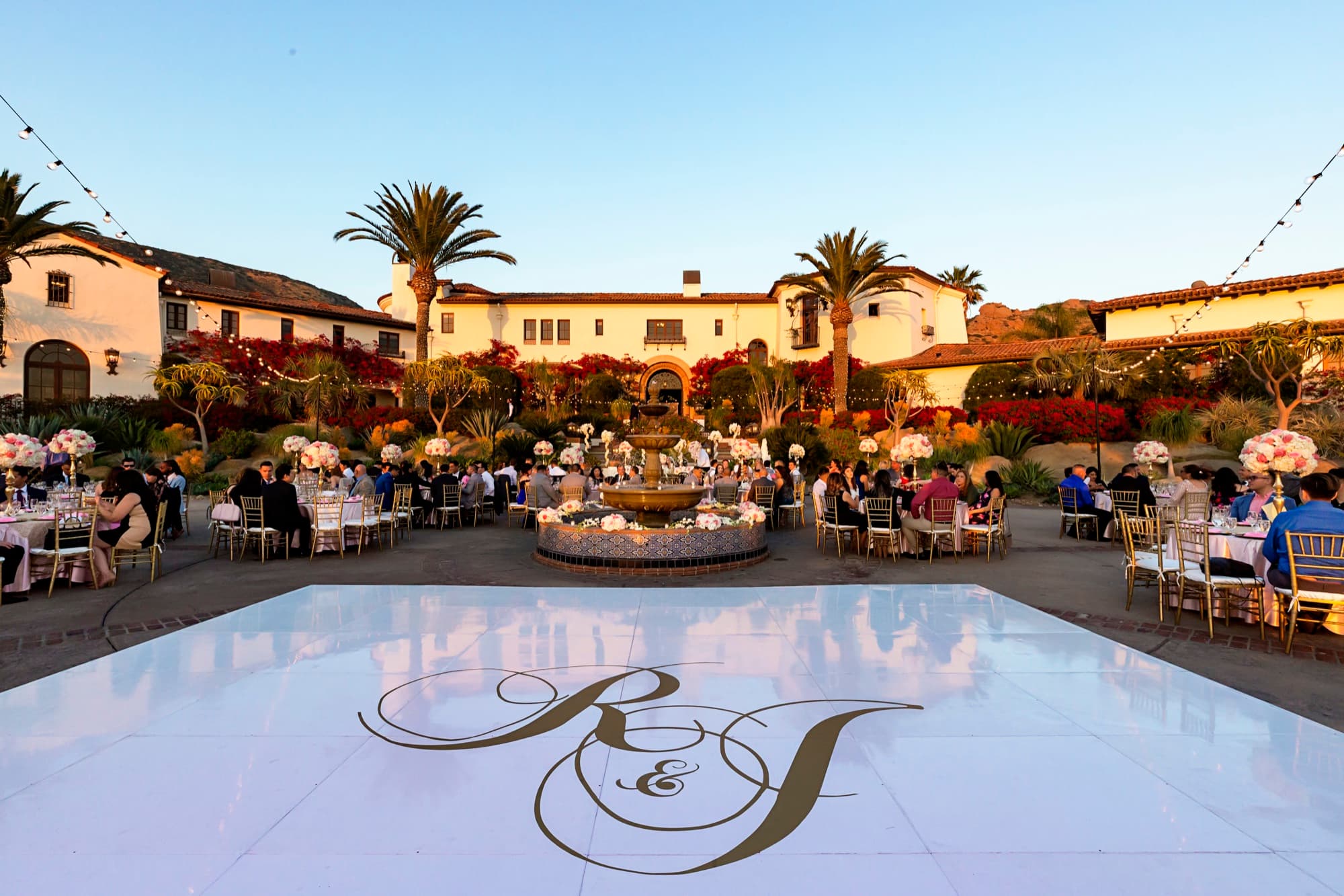 Courtyard reception at Hummingbird Nest Ranch with a monogrammed white dance floor, fountain centerpiece tables, and string lights at dusk