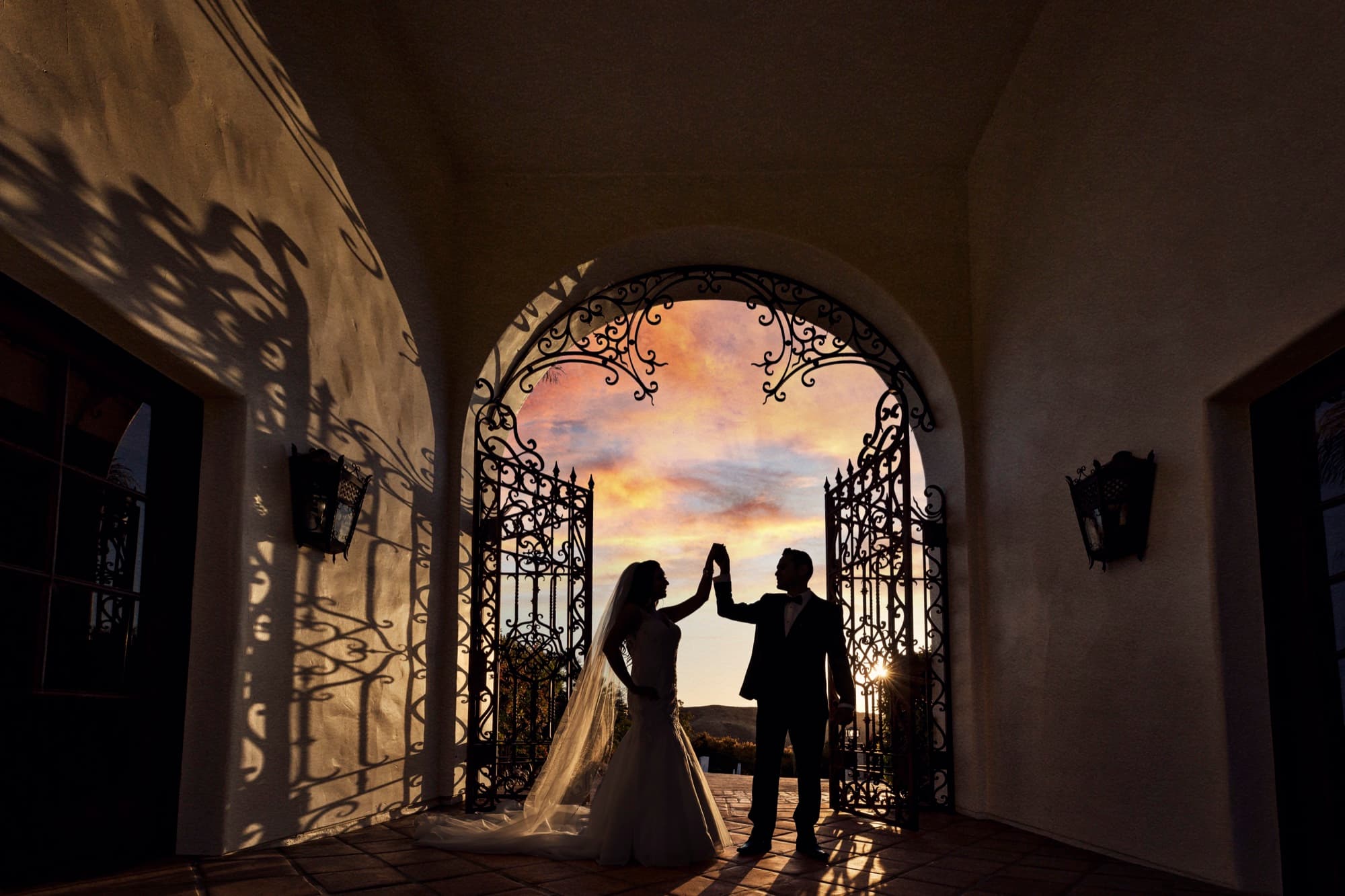 Bride and groom silhouetted in a high five against the sunset through the wrought-iron gates of Hummingbird Nest Ranch, photographed by Michael Anthony Photography