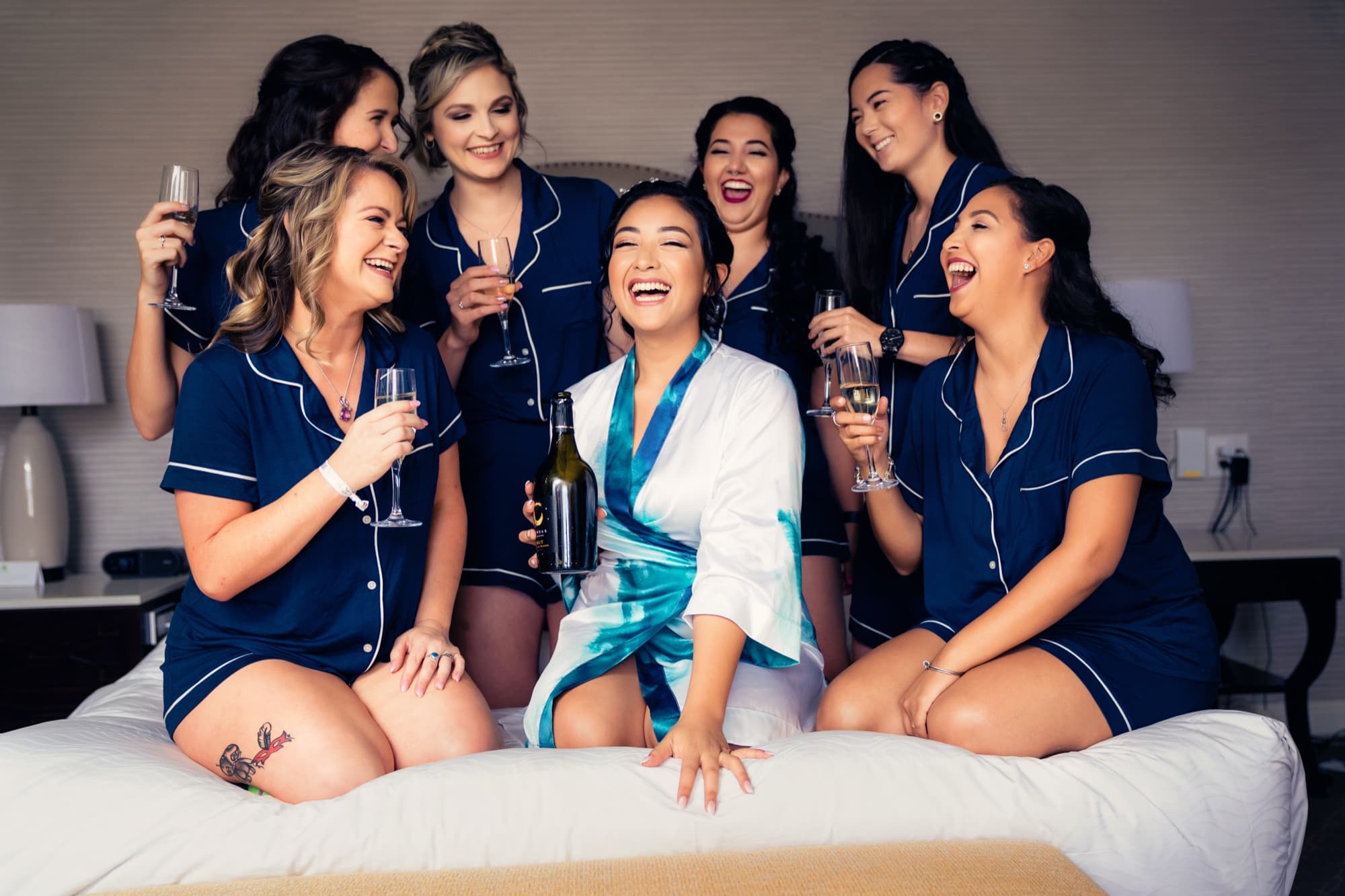 Ingrid in a white robe laughing with her bridesmaids in matching navy pajamas with champagne on the hotel bed