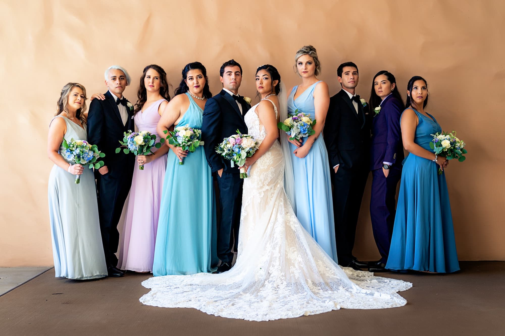Wedding party in mixed pastel gowns of periwinkle, lavender, and sky blue around Ingrid and her fanned lace train
