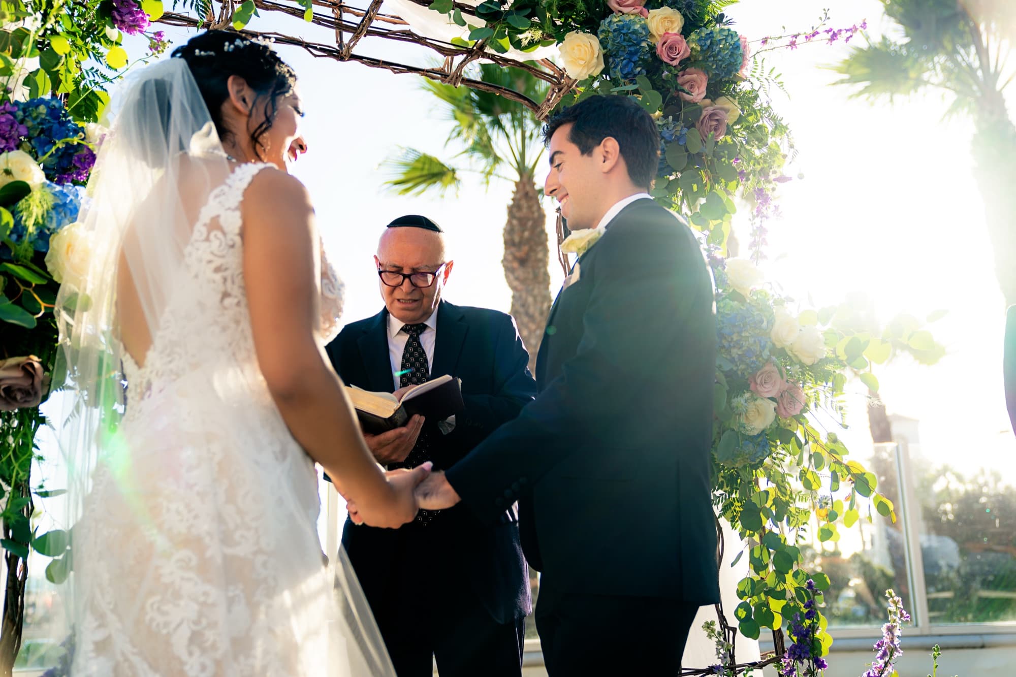 Ingrid and Navid holding hands during vows at the floral arch with sunlight flaring through the greenery
