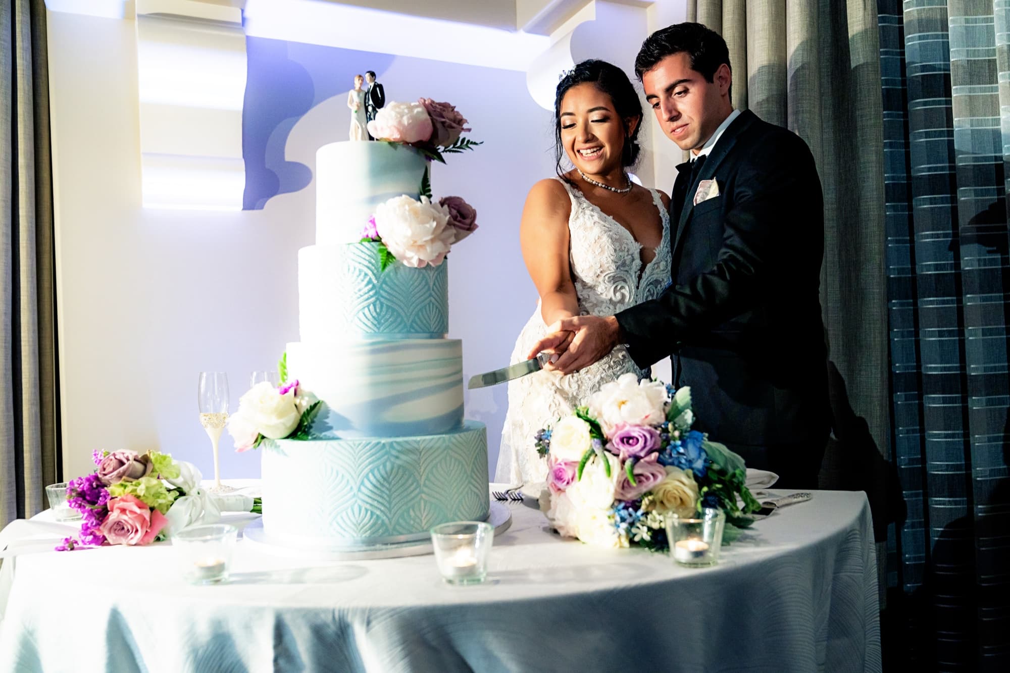 Ingrid and Navid cutting their teal and white tiered cake surrounded by purple and blue florals