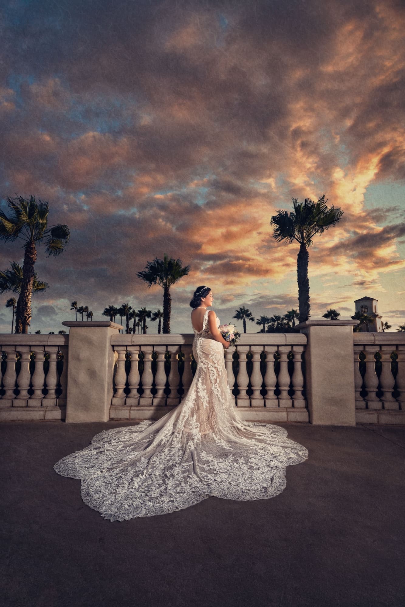 Ingrid on the stone terrace of the Hyatt Regency Huntington Beach at sunset, her lace cathedral train scalloped across the ground beneath palms and a burning sky, photographed by Michael Anthony Photography