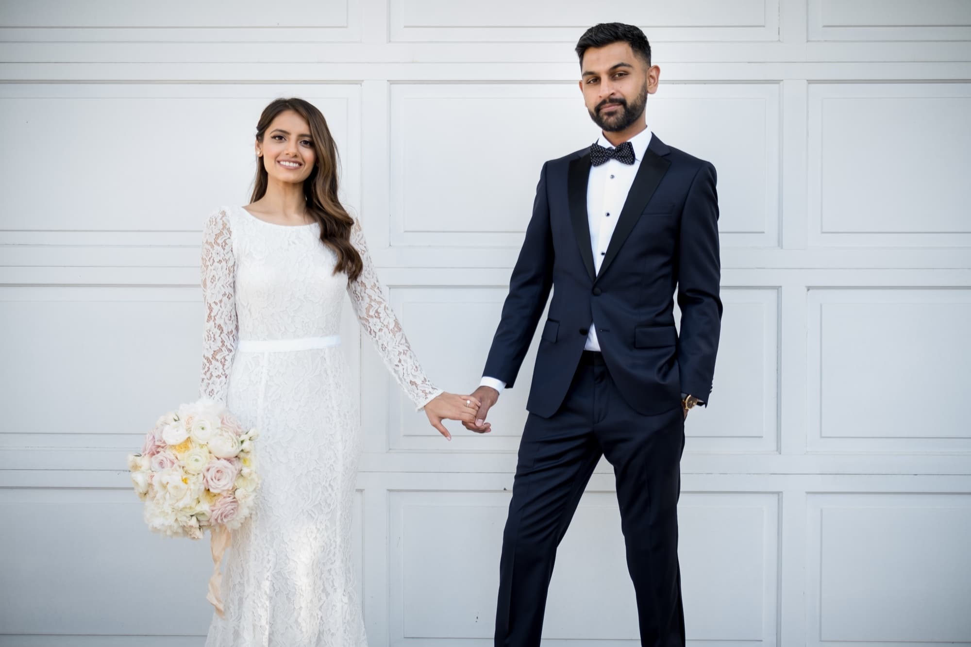 Formal portrait of Noor in her long-sleeve lace gown and Sheazad in his tuxedo holding hands against a white paneled wall