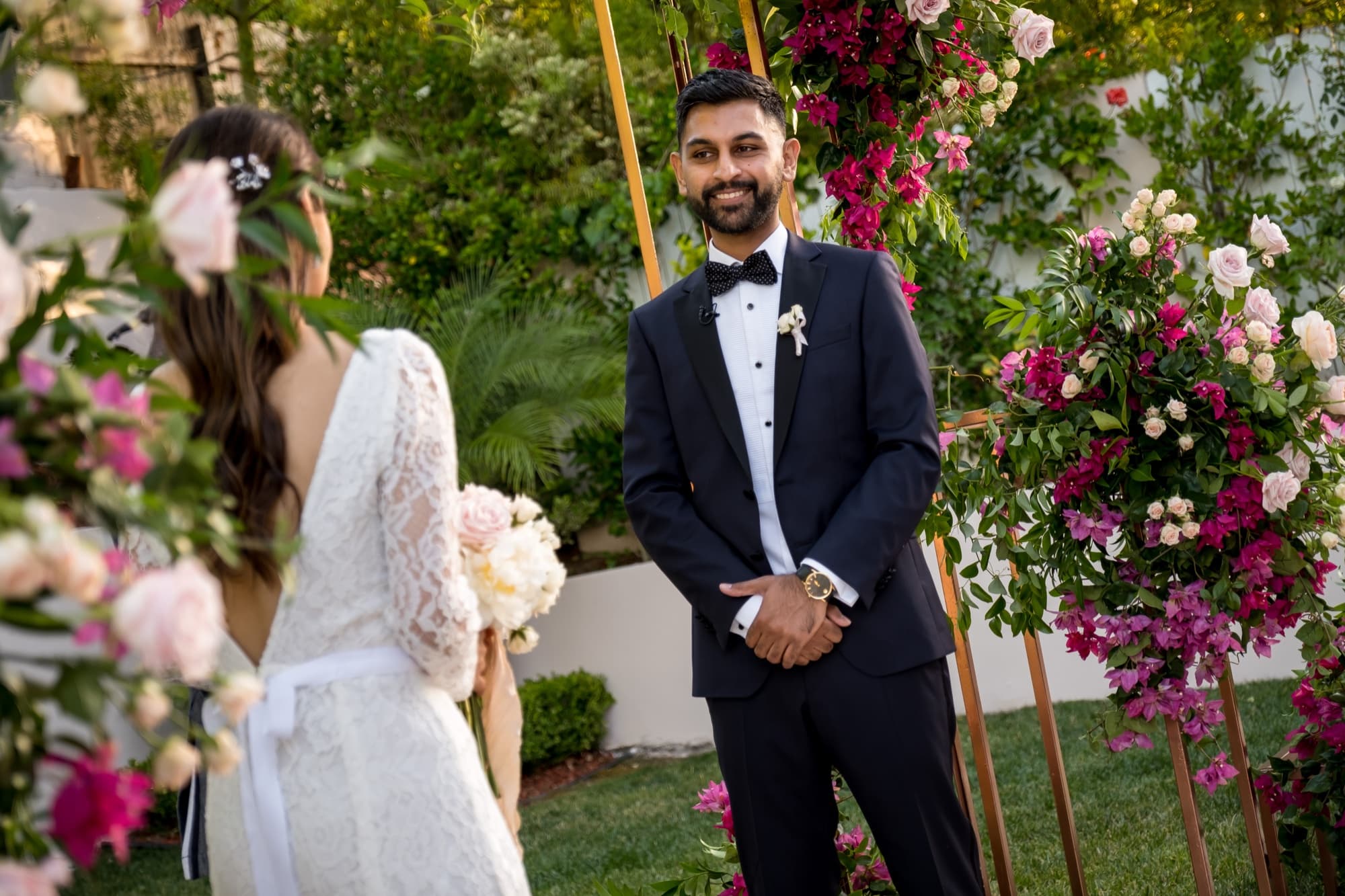 Sheazad reading his vows from a folded page to Noor beneath the rose and bougainvillea arch