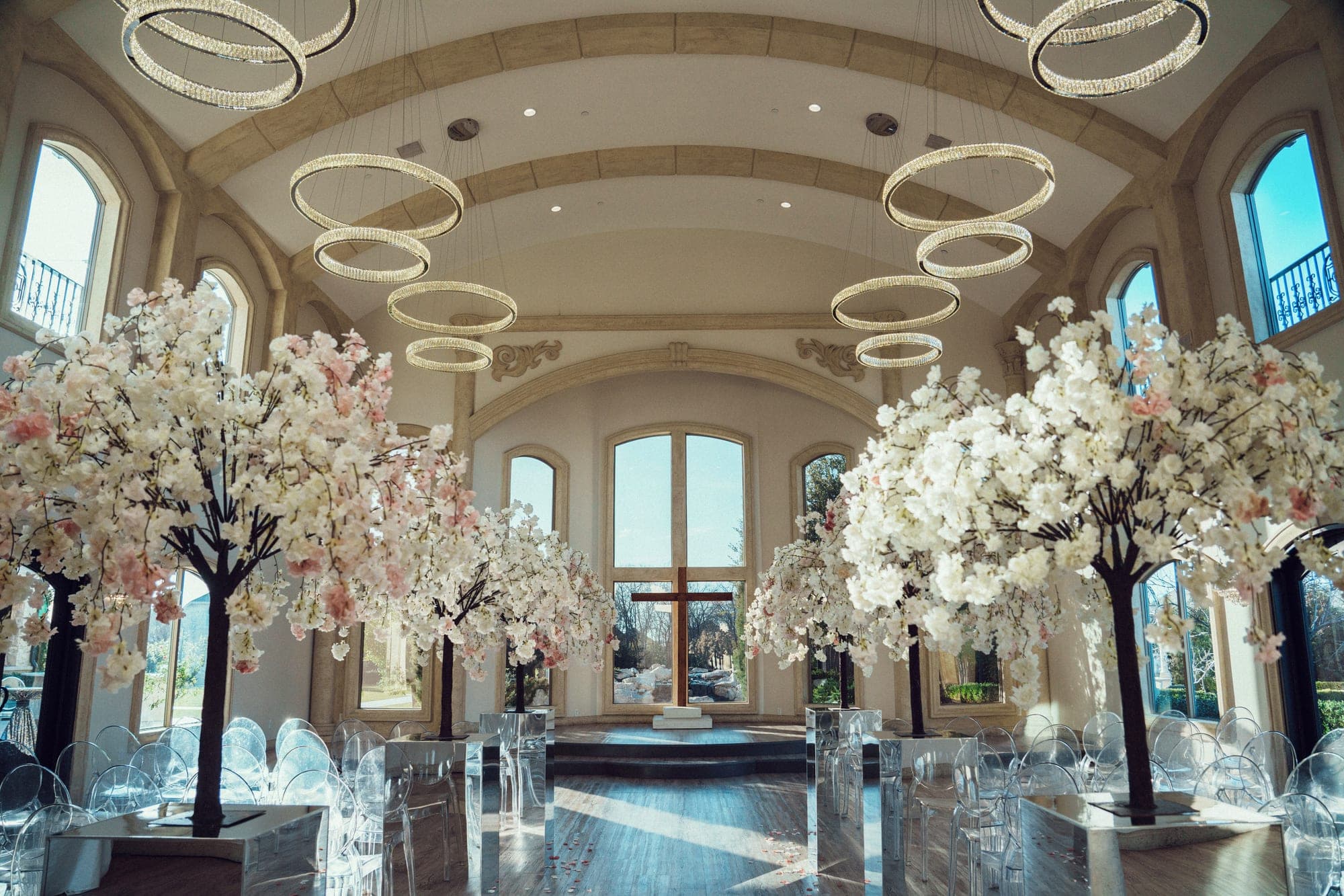The glass chapel at Knotting Hill Place set with white and blush blossom trees, ghost chairs, and halo chandeliers