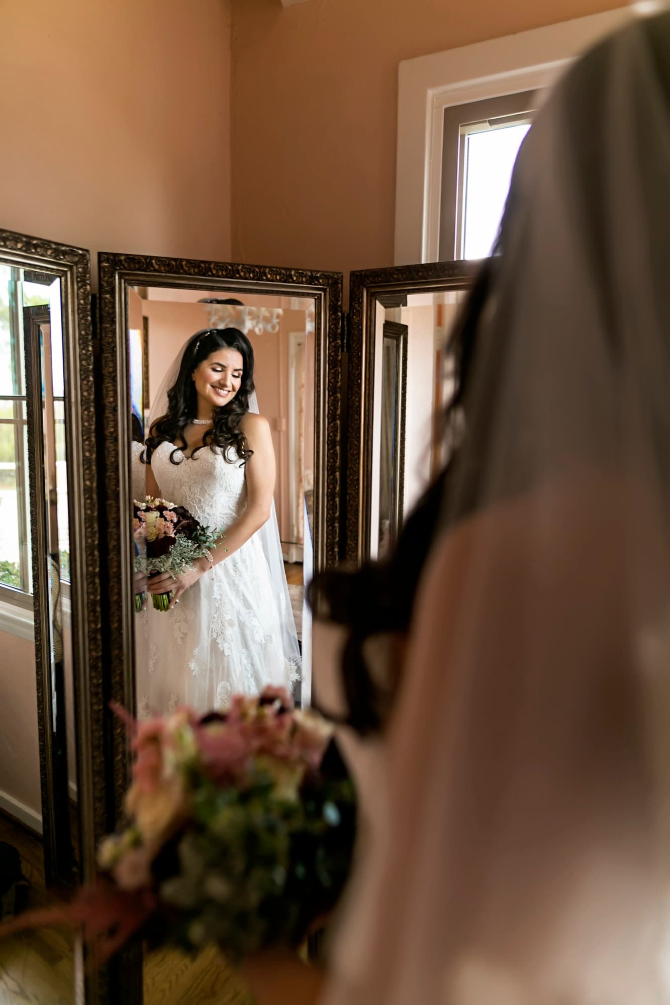 Stephanie in her lace gown smiling at her reflection in the antique mirror at La Venta Inn while holding a burgundy bouquet