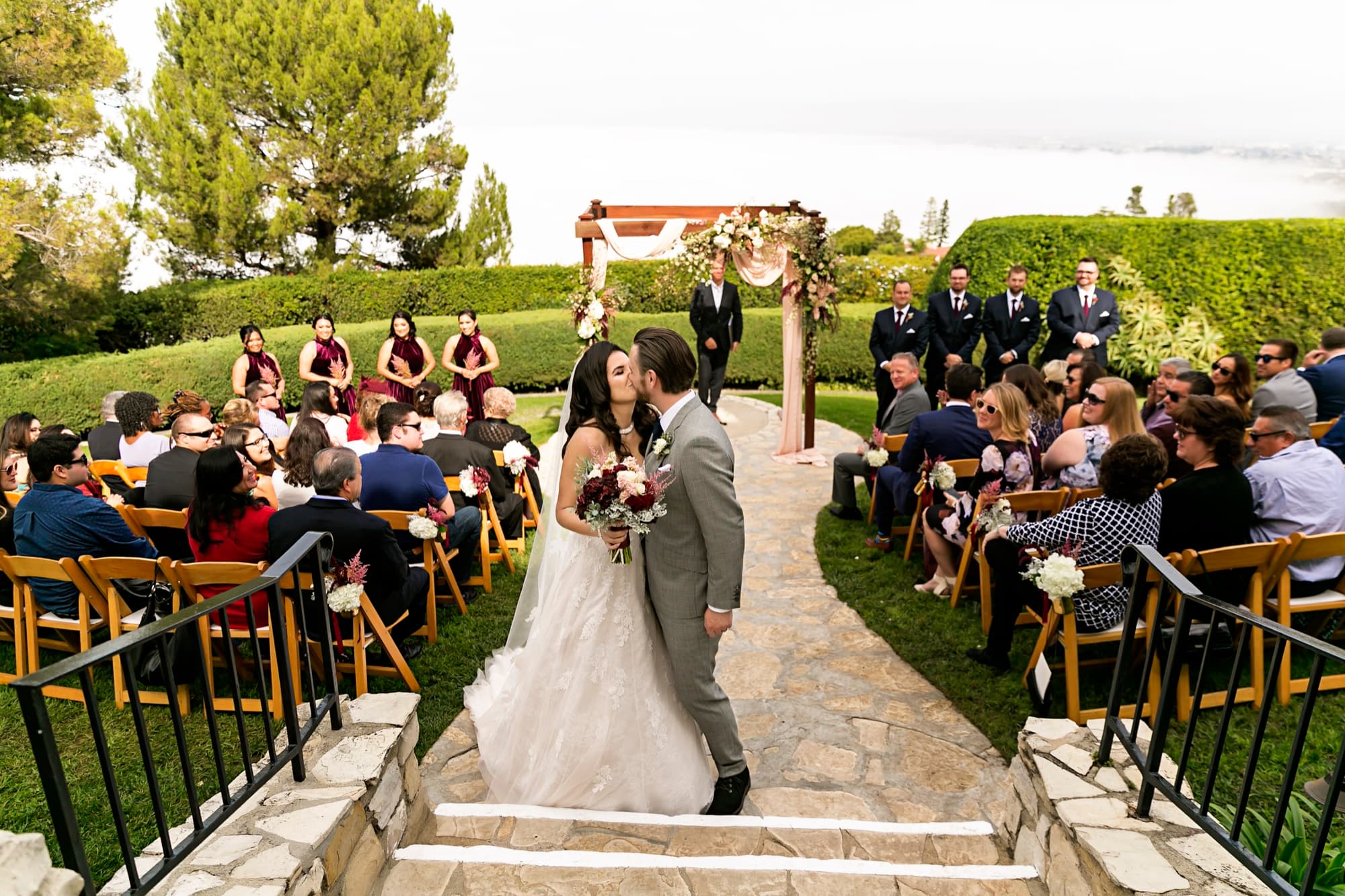 Stephanie and Josh kissing on the stone aisle after their ceremony as guests cheer on the ocean-view terrace at La Venta Inn