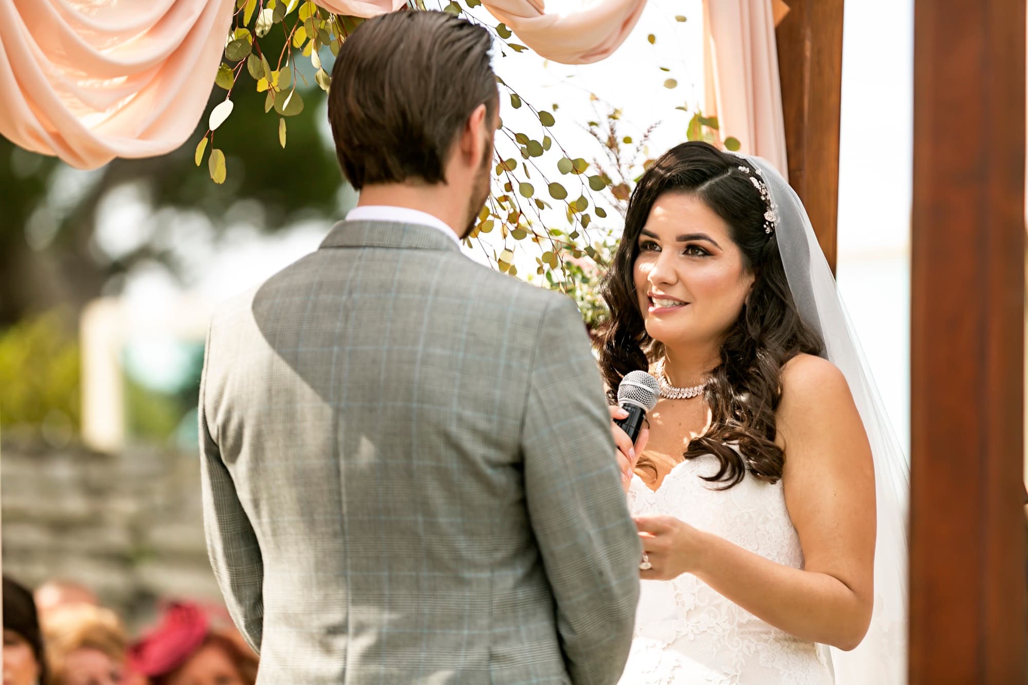 Stephanie speaking her vows into a microphone under the blush-draped arch as Josh listens
