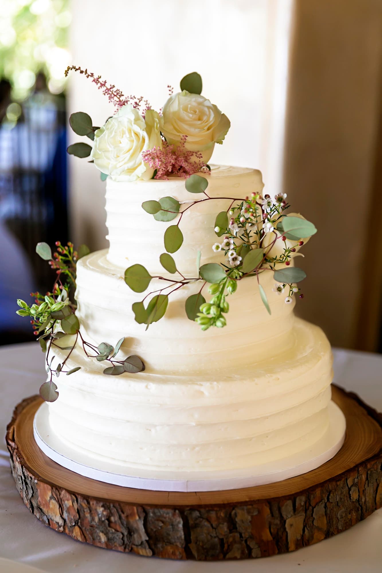 Three-tier buttercream wedding cake with white roses and eucalyptus on a wood slab at La Venta Inn