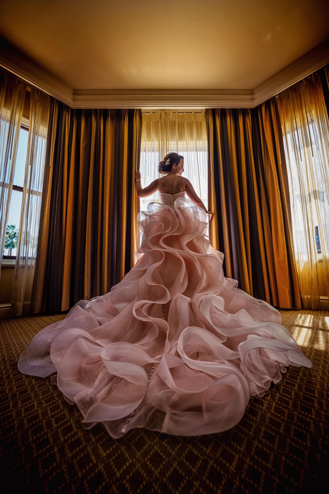 Ilene standing at the corner window of her Langham suite with the blush ruffled train of her ballgown spread across the floor