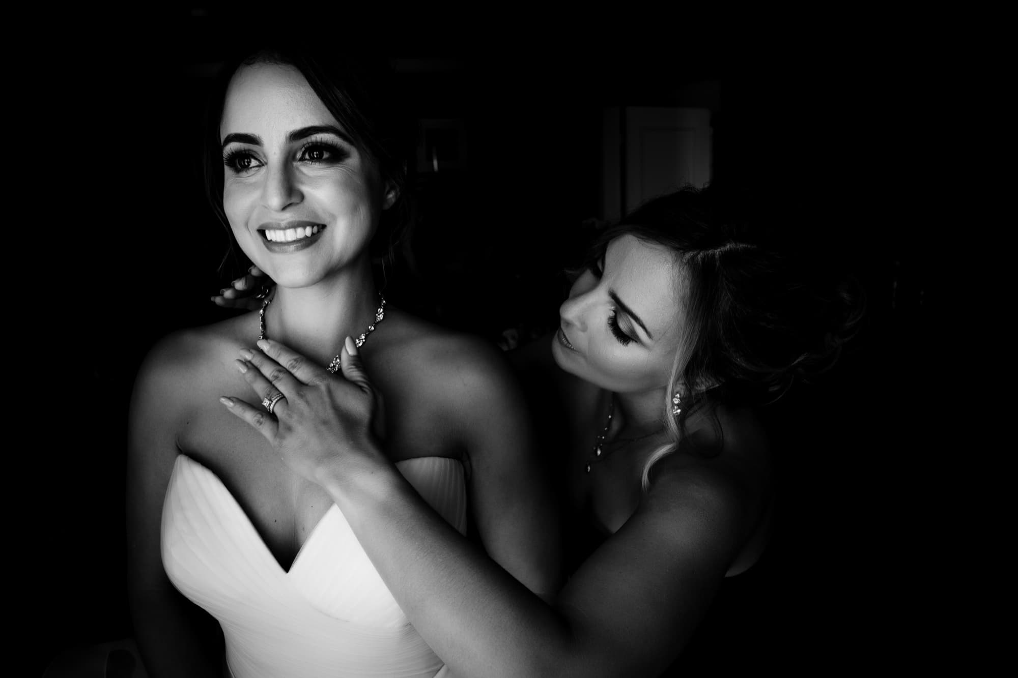 Black and white photograph of the bride's sister fastening her necklace before the ceremony