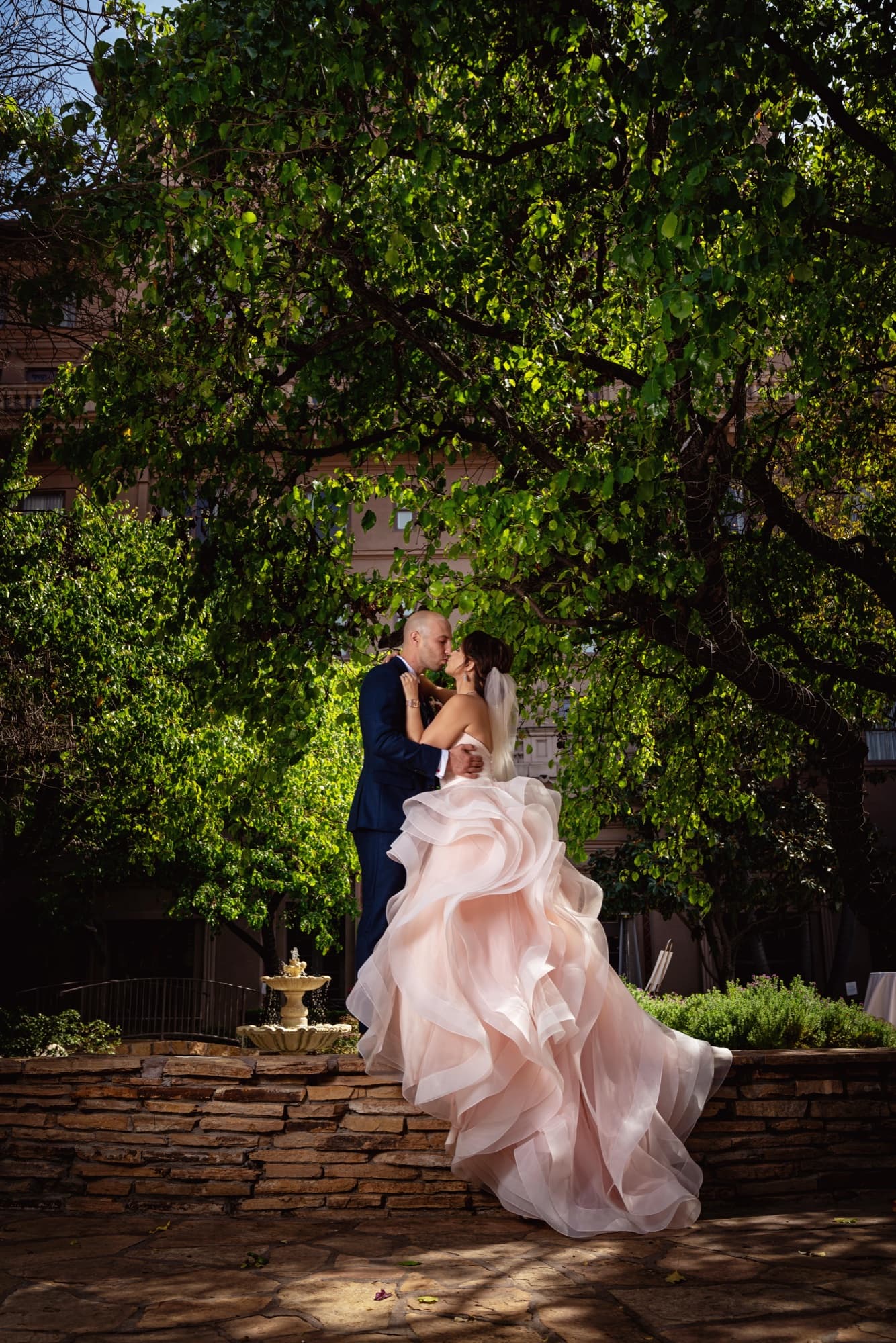 Ilene and Ricardo embracing beside the stone fountain under the trees at The Langham Huntington Pasadena