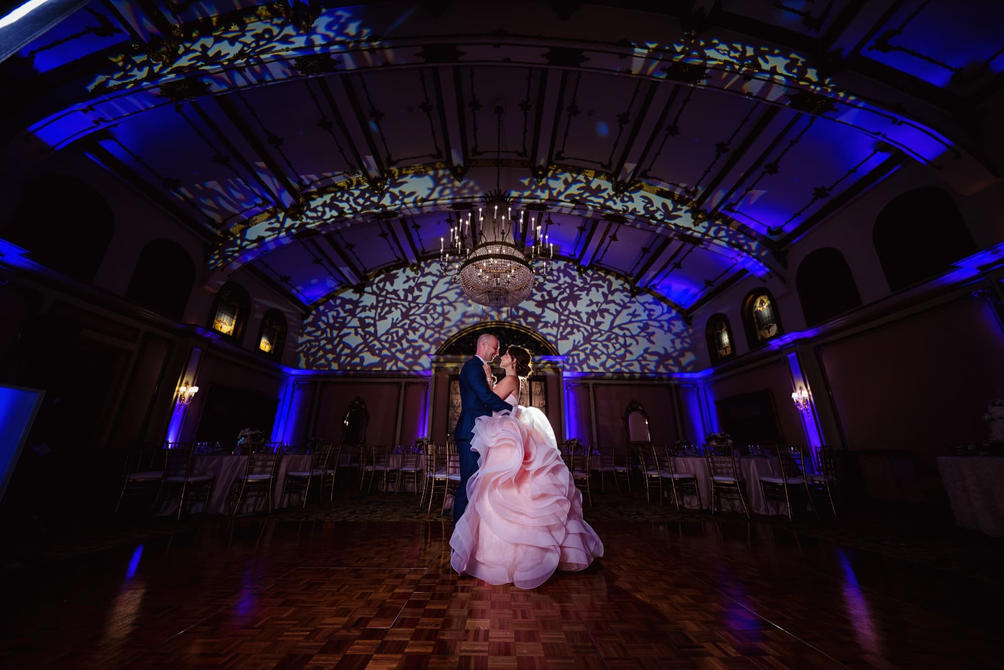 Ilene and Ricardo dancing alone on the parquet floor of the Langham Huntington ballroom under a chandelier and patterned ceiling light