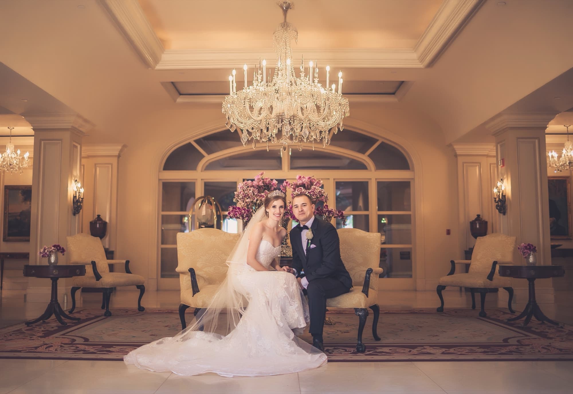 Ilana and Christopher seated on a settee beneath a crystal chandelier at the Langham, her veil cascading across the floor