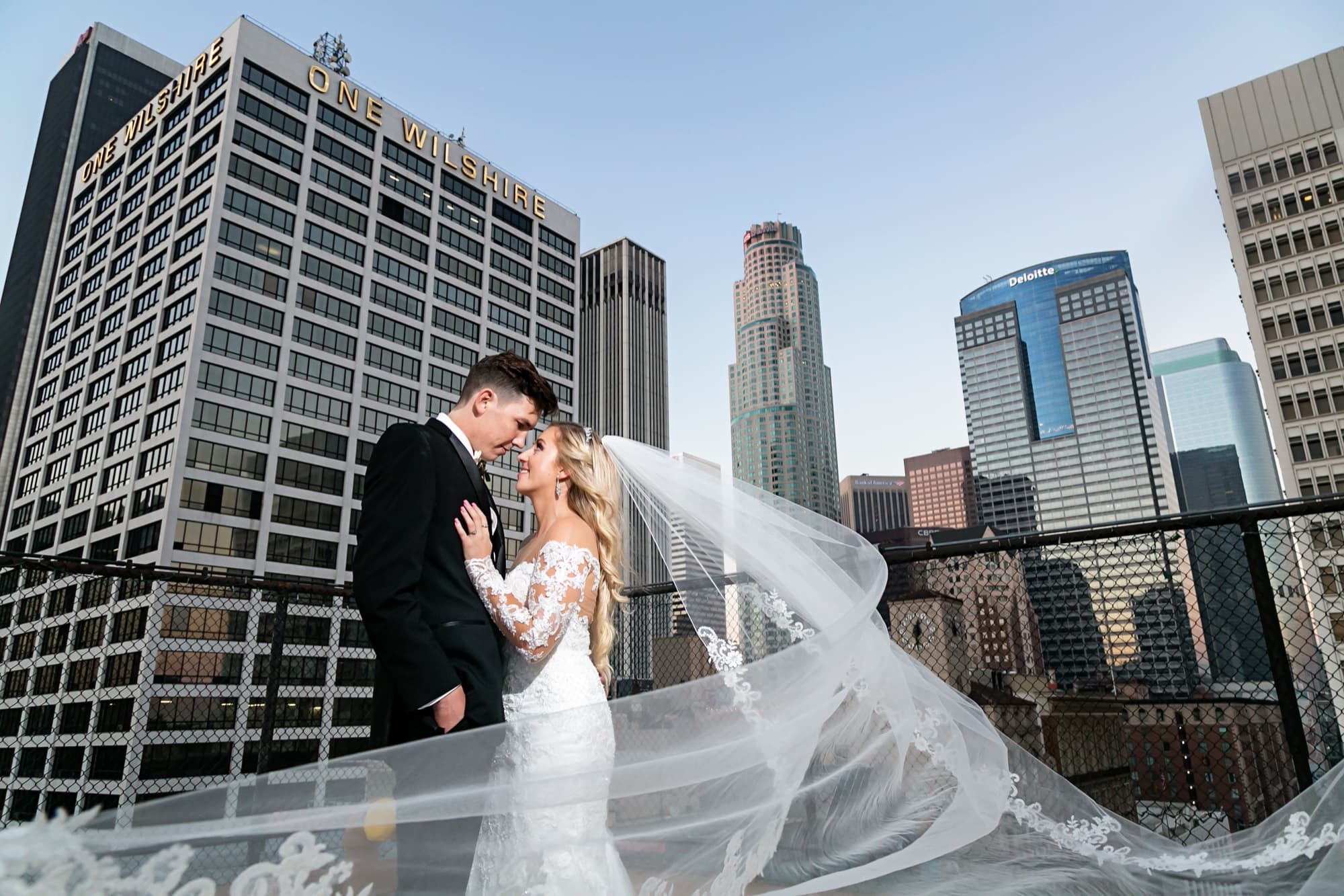 Jaymie and Matt embracing on the Los Angeles Athletic Club rooftop with her cathedral veil streaming across the downtown skyline, photographed by Michael Anthony Photography