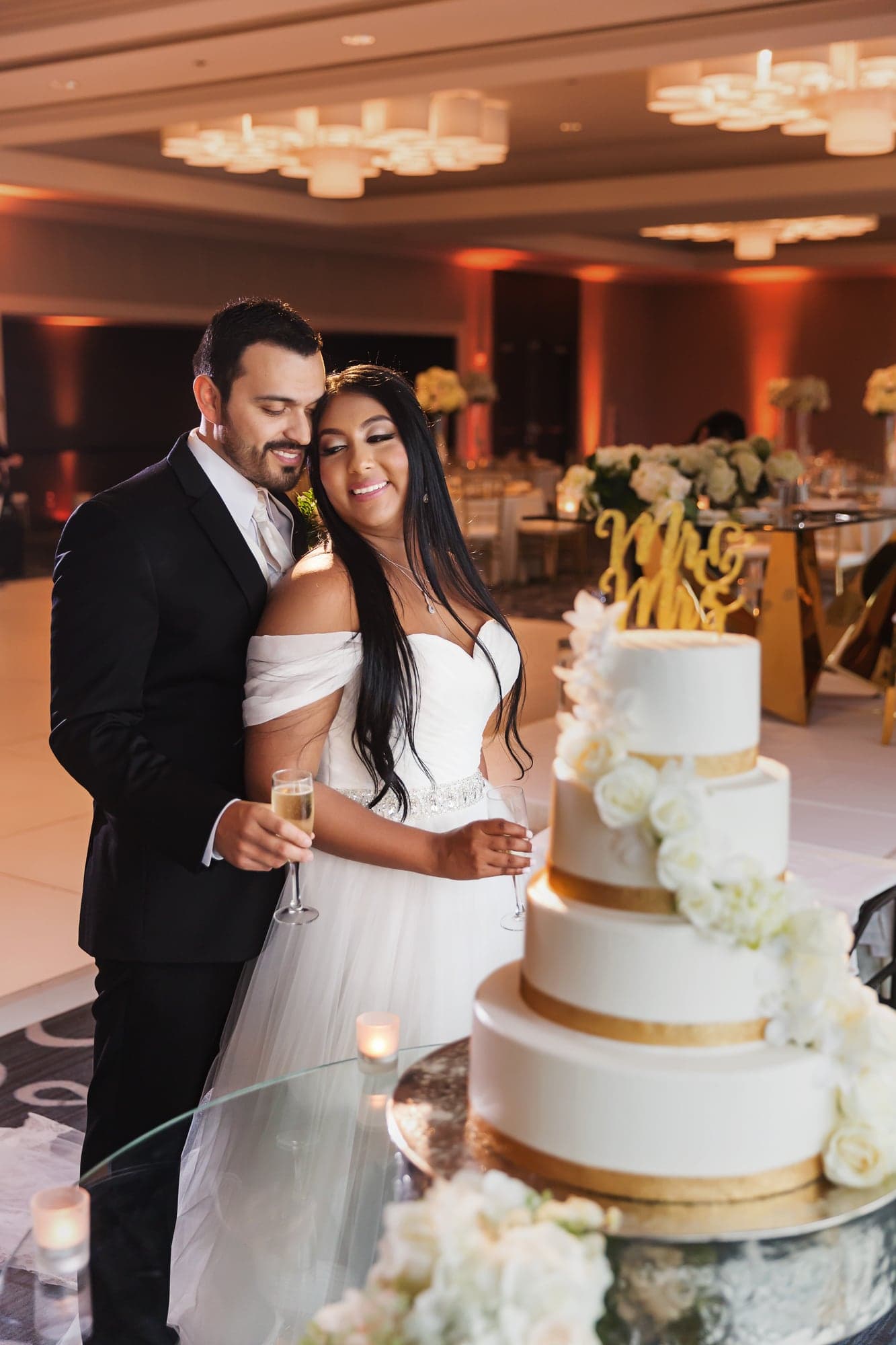 Tari and JJ standing together beside their white-and-gold wedding cake