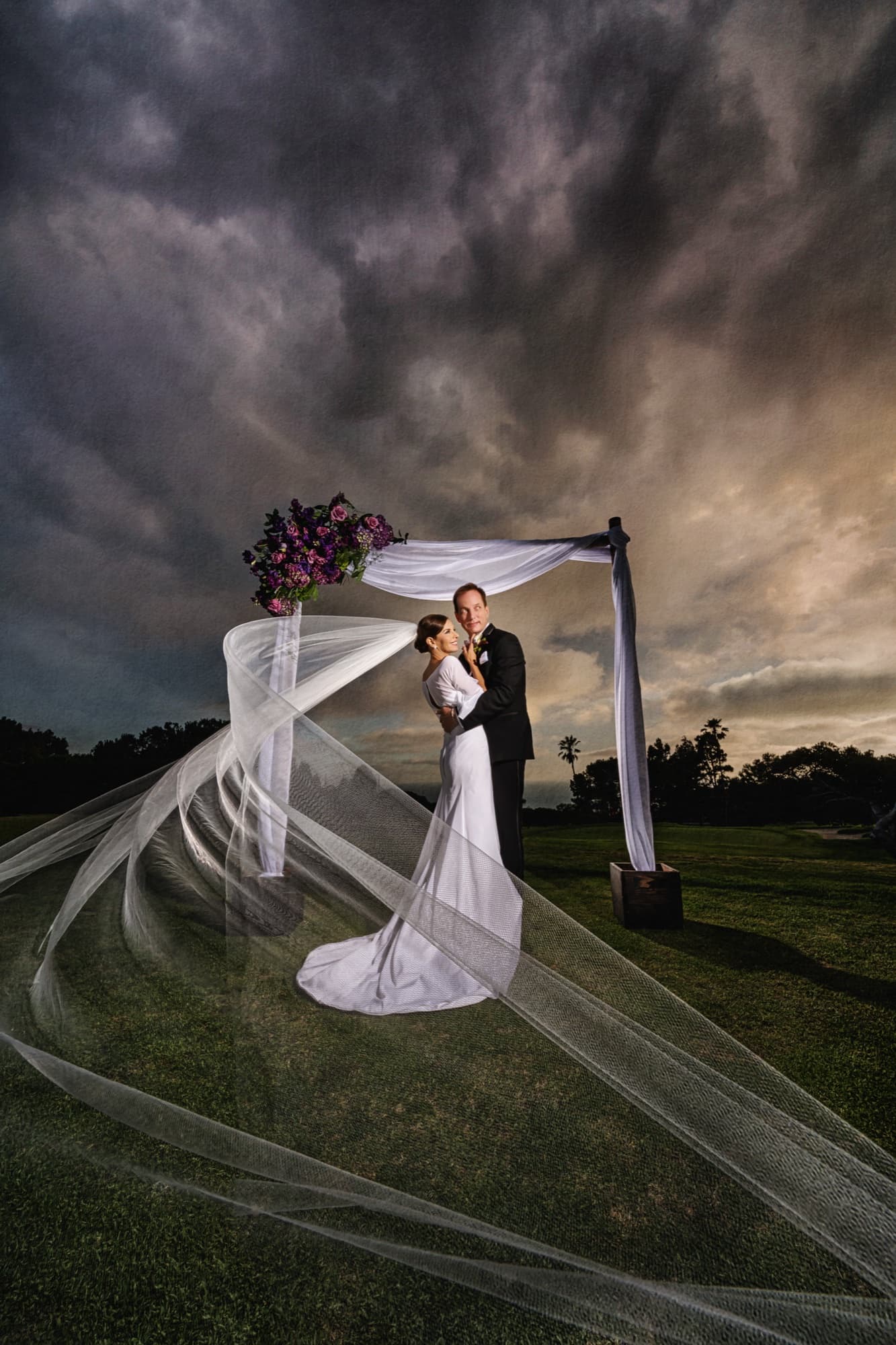 Andrea's cathedral veil sweeping in the wind at the draped ceremony arch under dramatic storm clouds at Los Verdes Golf Course