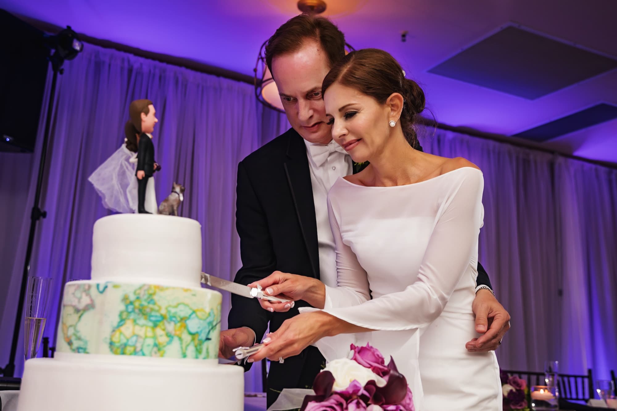Andrea and John cutting their travel-themed wedding cake with a painted world map tier and figurine topper with their cat