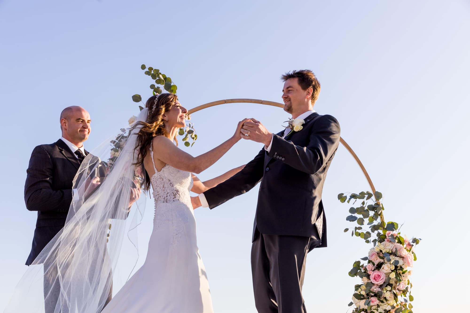 Arta and her groom joining hands beneath a circular floral arch on the sand during their Malibu beach ceremony