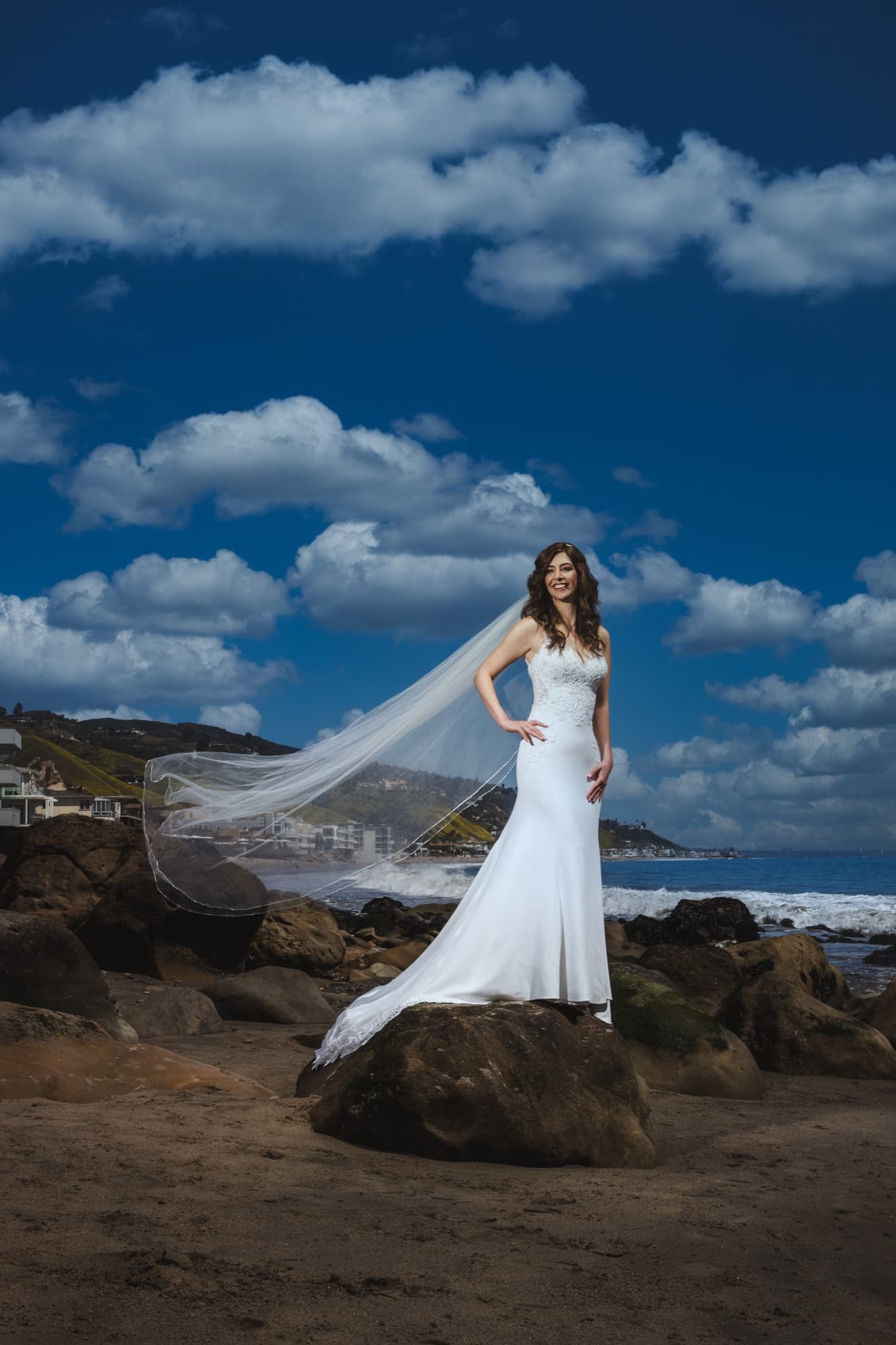 Arta standing on a boulder at the Malibu tideline in her lace gown, veil trailing, against a bright blue sky and clouds