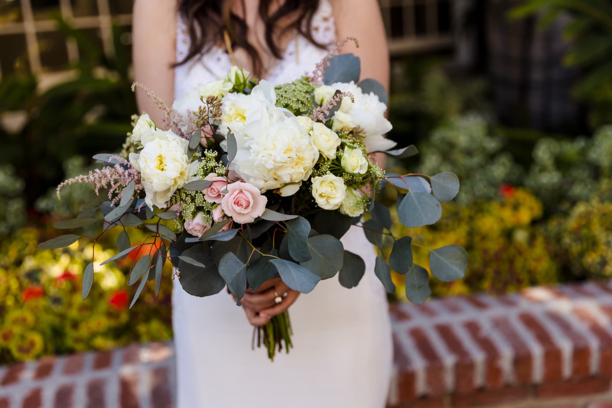 A loose garden-style bouquet of ivory roses, blush, and eucalyptus held against a brick planter at a Malibu wedding