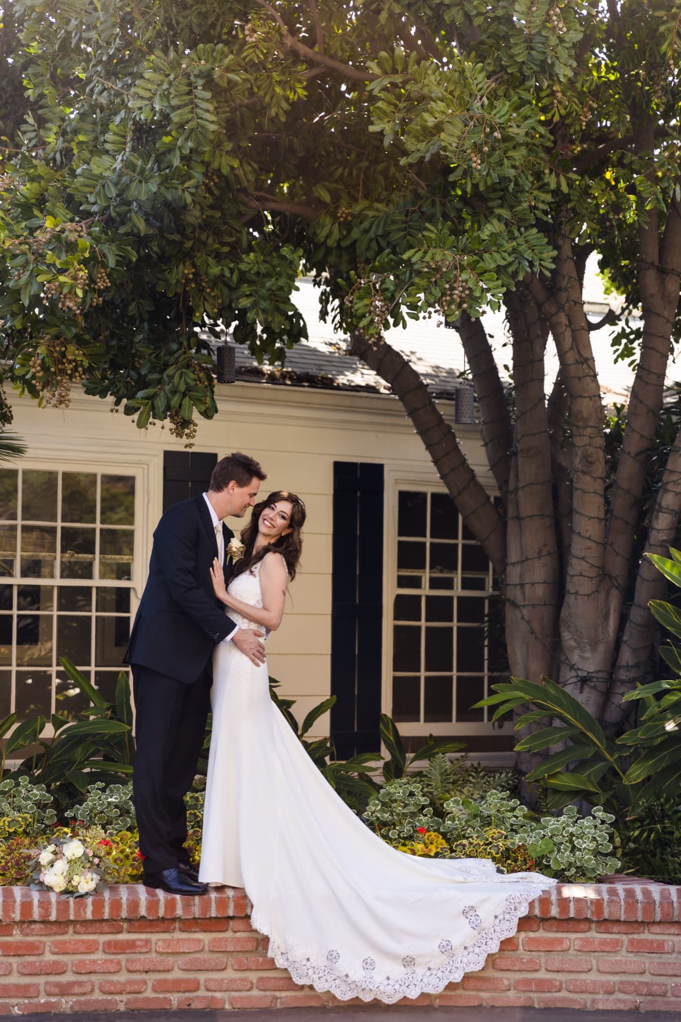 Arta and her groom embracing in the garden of a Malibu beach cottage, her train spilling over a brick wall