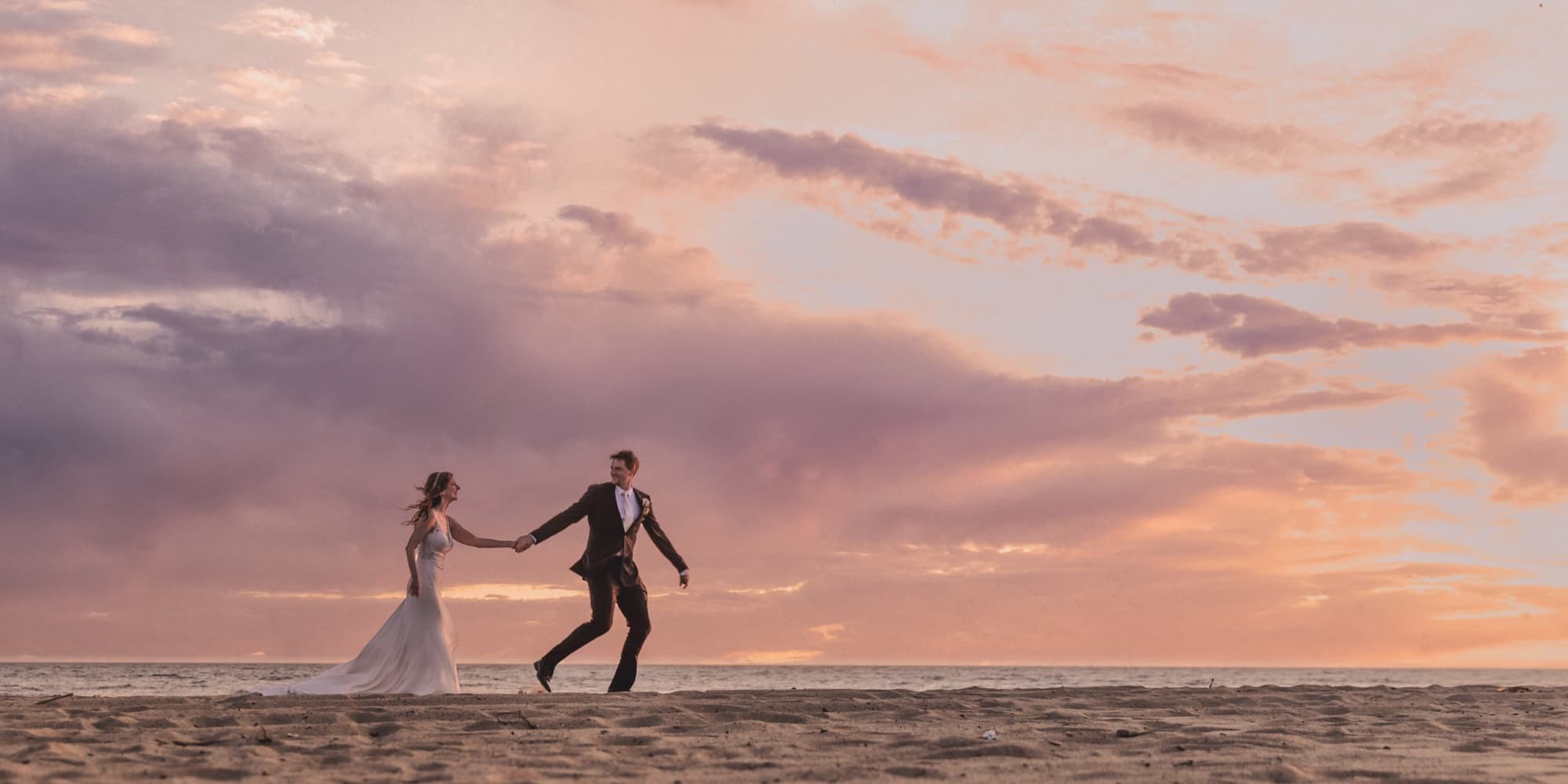 Arta and her groom playing on the Malibu sand at sunset, hands linked, under a wide pink and lavender sky, photographed by Michael Anthony Photography