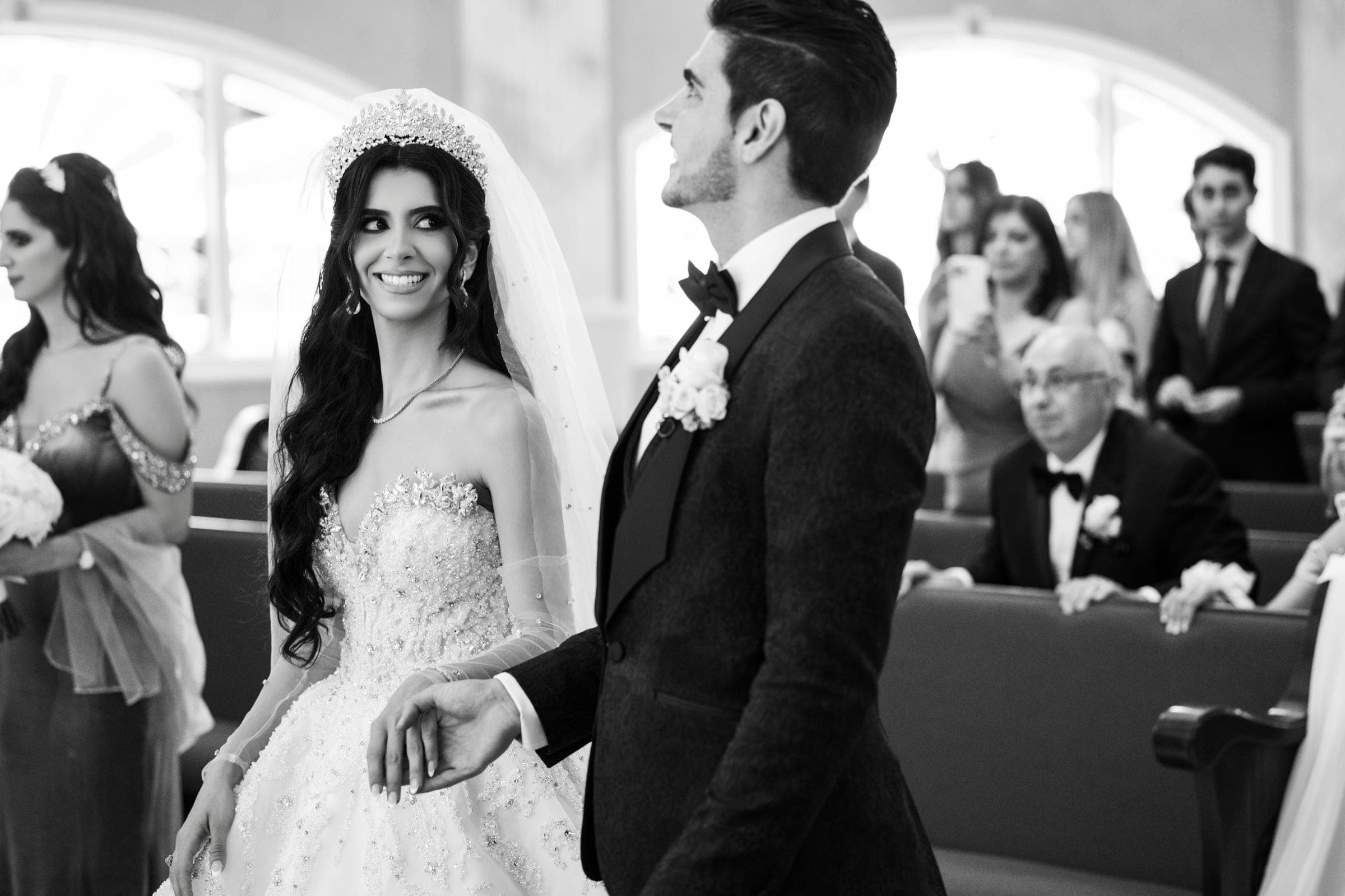Smiling bride in tiara and veil holds her groom's arm inside the church as guests watch