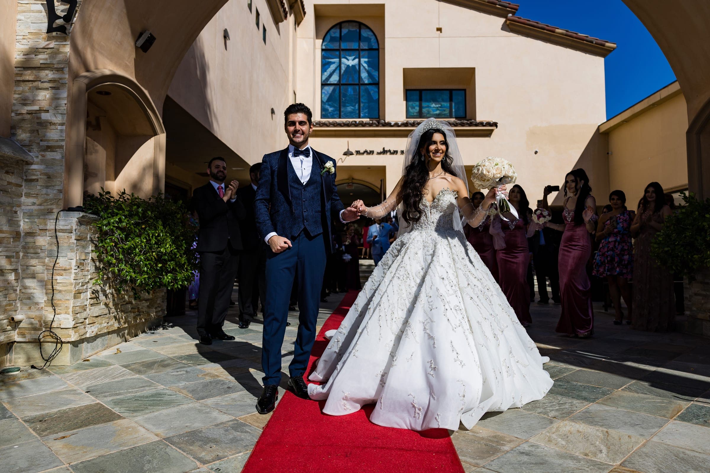 Groom in blue suit leads bride down a red carpet outside as guests applaud in a sunny courtyard