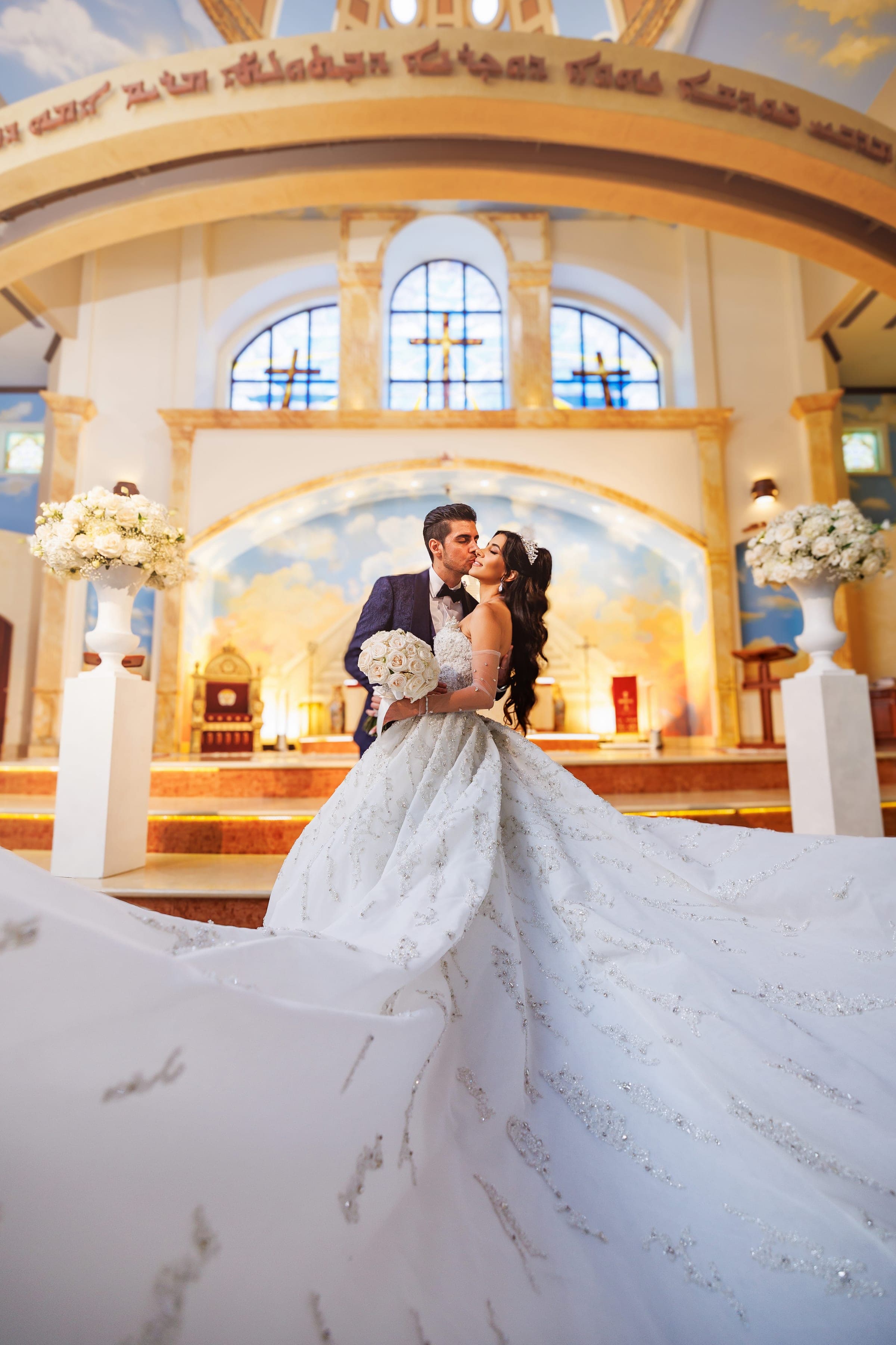 Groom kisses bride's cheek at the altar as her long beaded train sweeps toward the camera