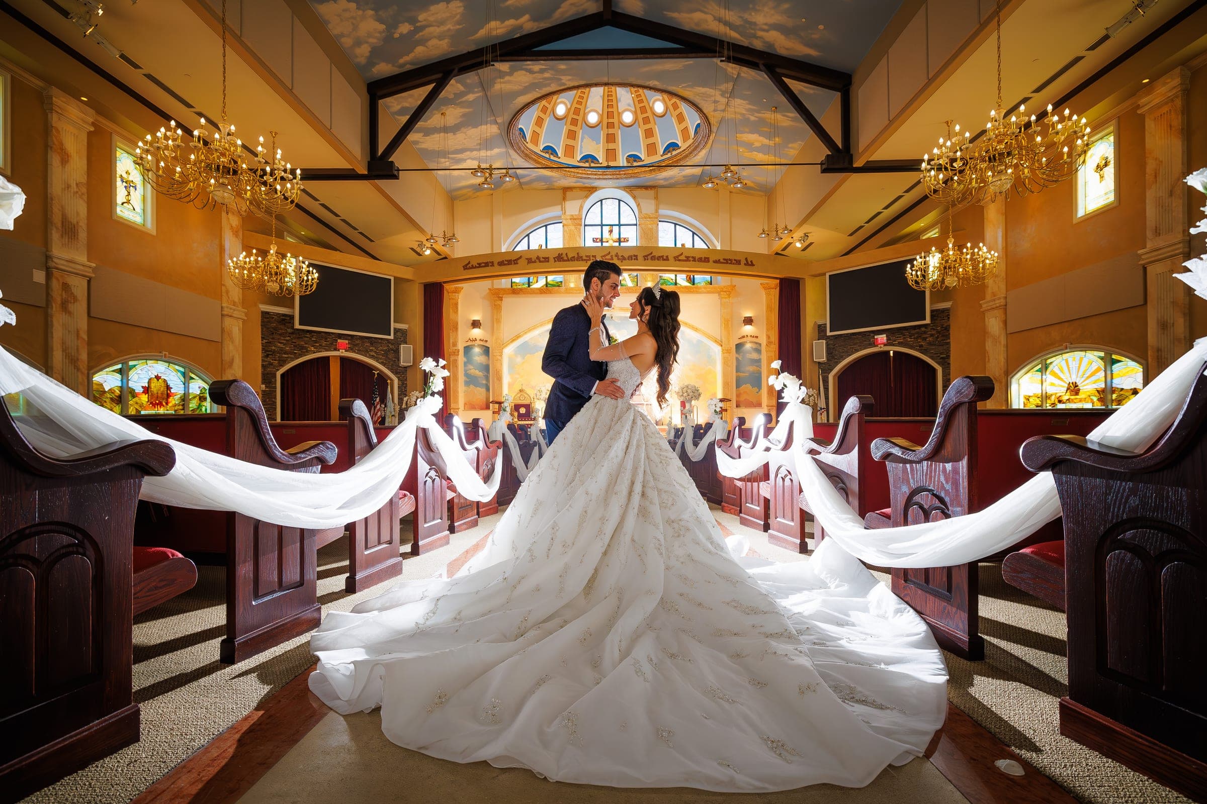 Groom holds bride close between draped pews, her voluminous gown spread across the church floor