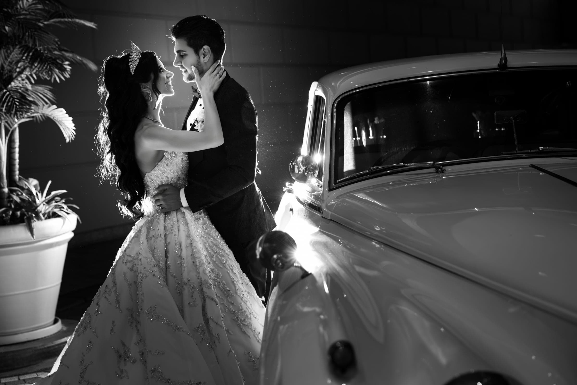 Dramatic black and white portrait of the couple embracing beside a vintage white car at night