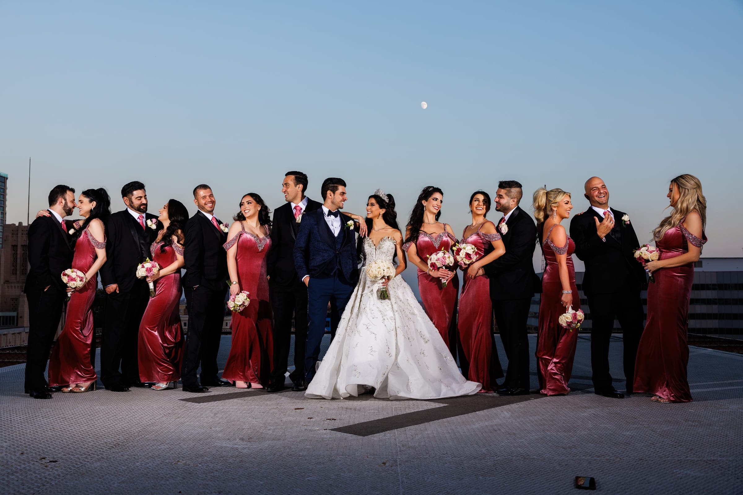 Wedding party in burgundy and dark suits lines up beside the couple on a rooftop at dusk