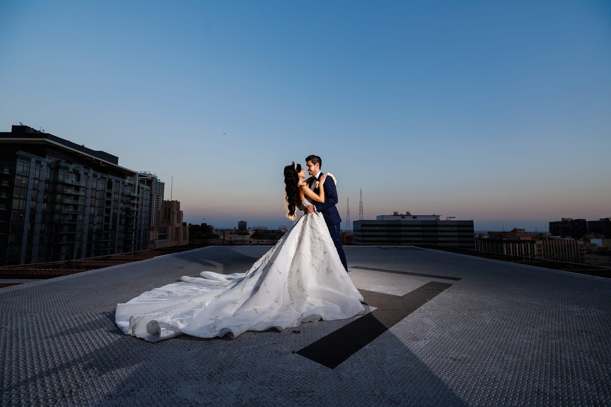 Bride and groom kiss on a rooftop at dusk, her long gown train sweeping the concrete against city buildings.
