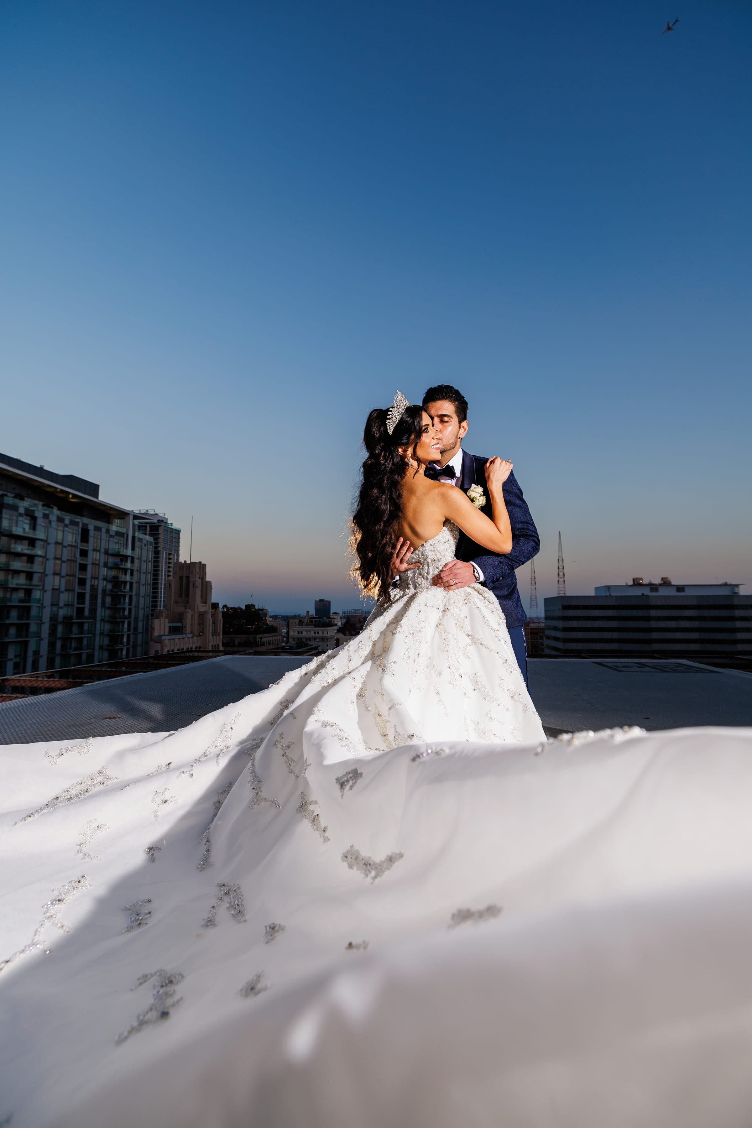 Couple embraces closely on a rooftop as the bride's beaded gown train billows dramatically across the ground.