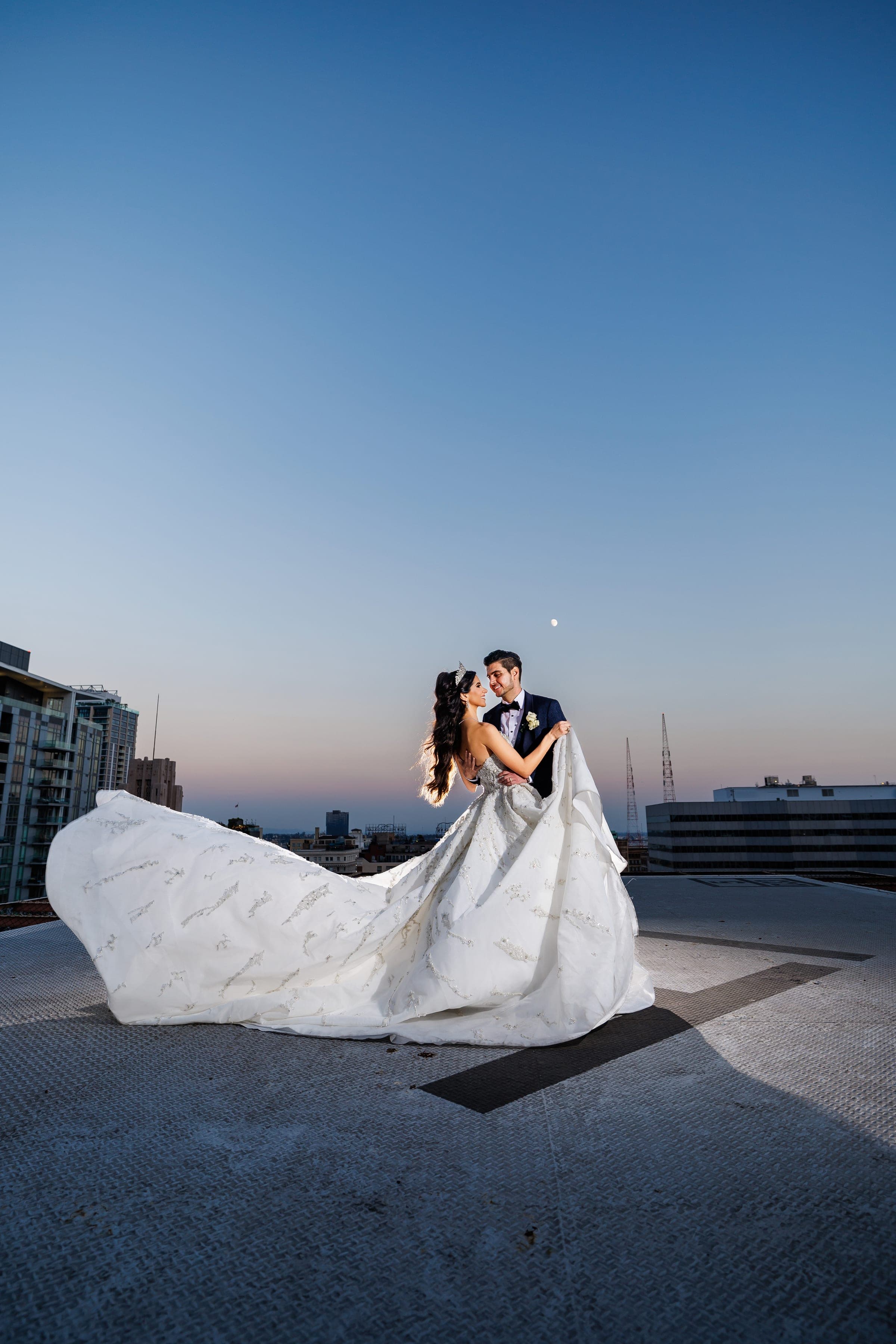 Bride and groom stand facing each other on a rooftop, her train fanned wide under a moonlit twilight sky.
