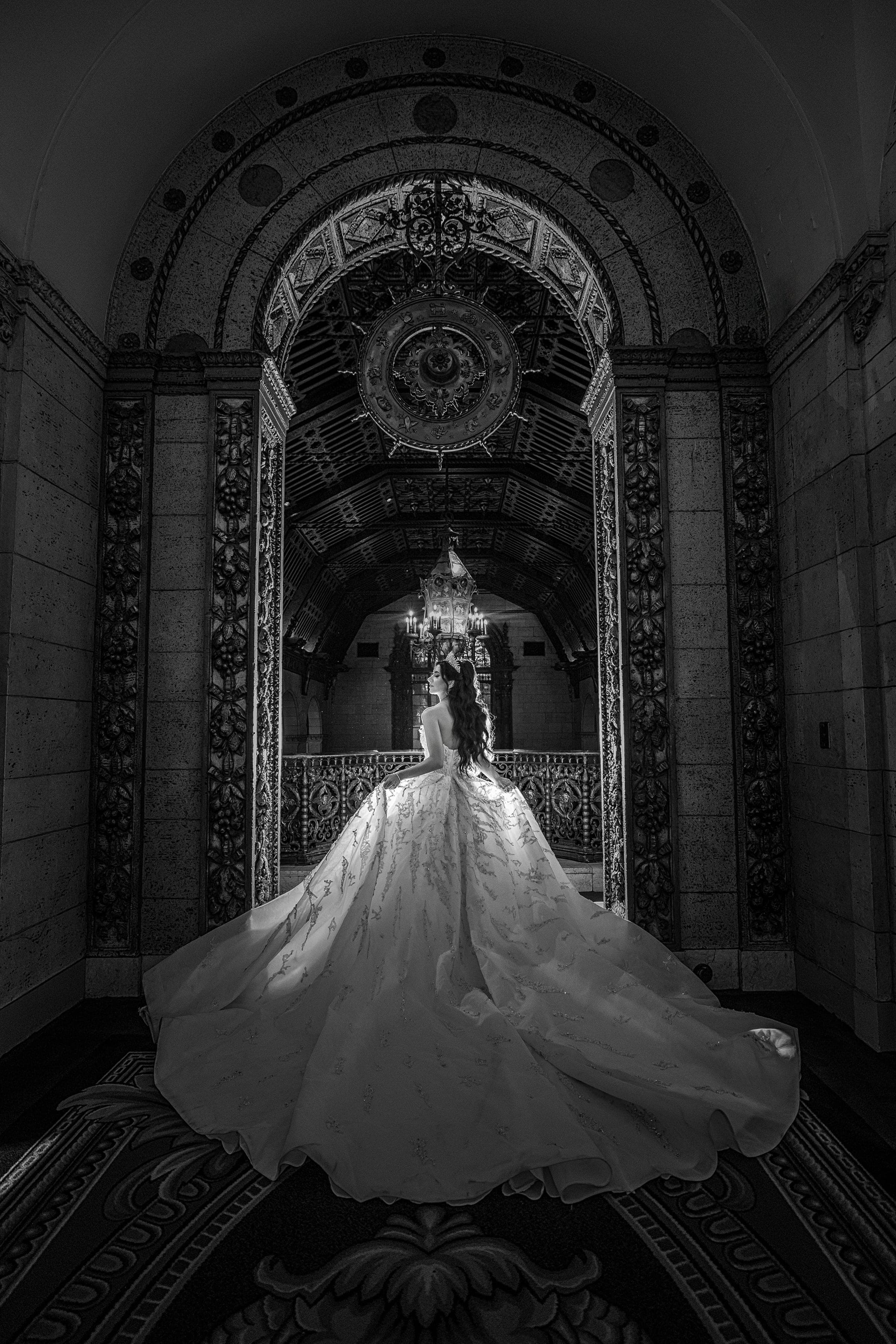 Black and white image of a bride facing an ornate arched hallway, her voluminous gown spread across patterned carpet.