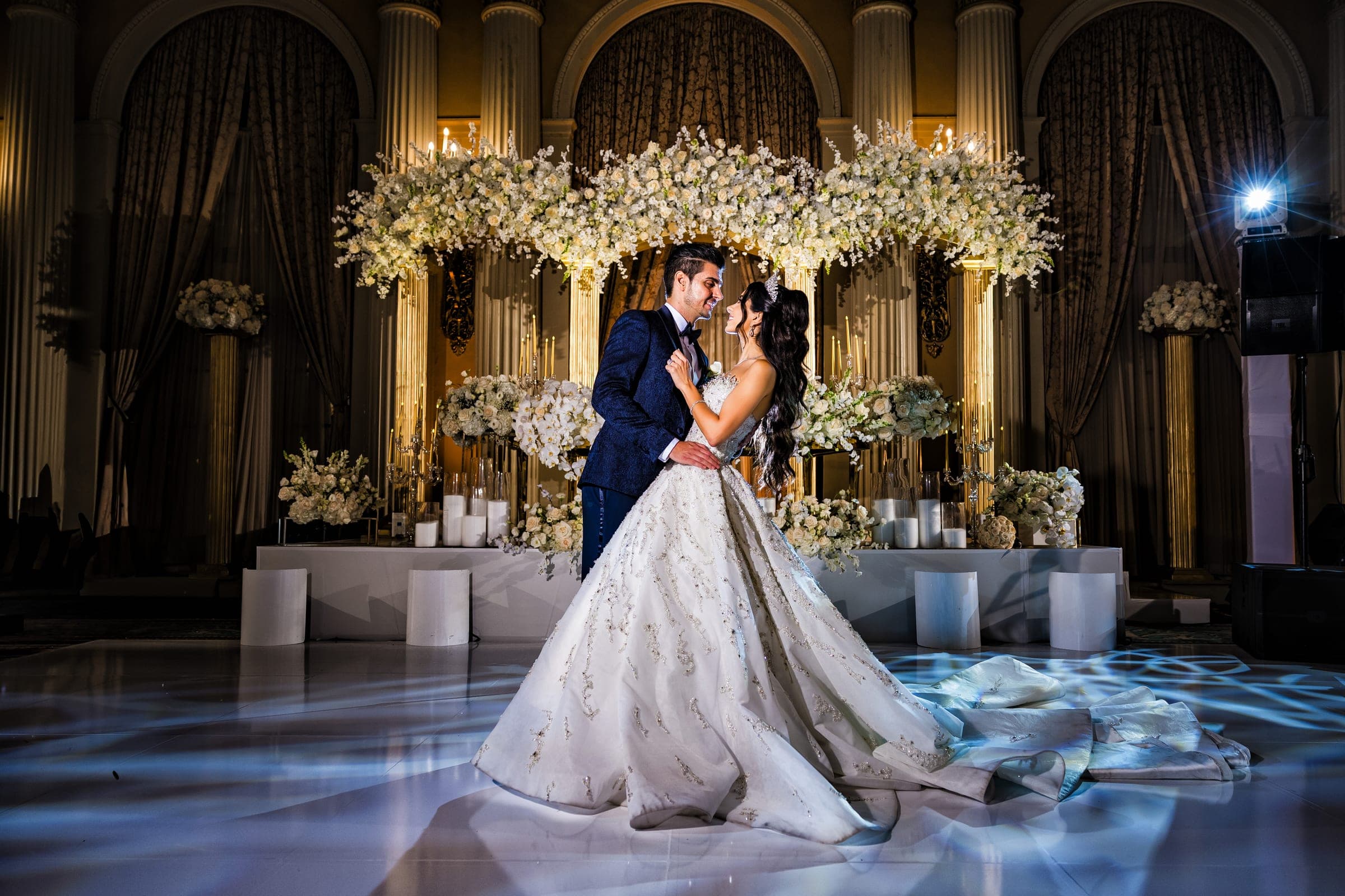Couple embraces beneath a lush white floral arch on an illuminated ballroom dance floor with candles and columns.