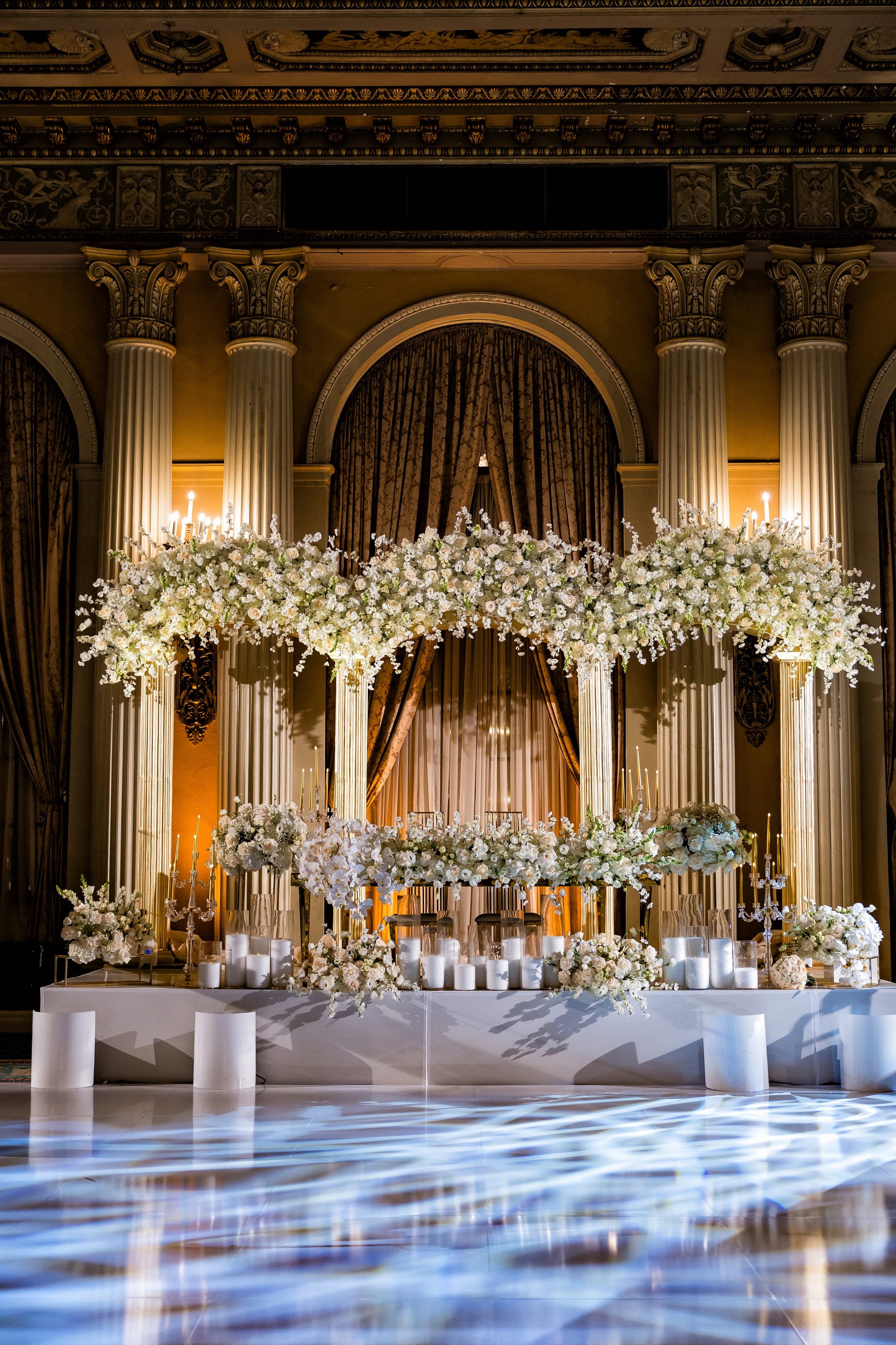 Elaborate white floral arch and candle display on a raised stage before tall columns and draped ballroom walls.