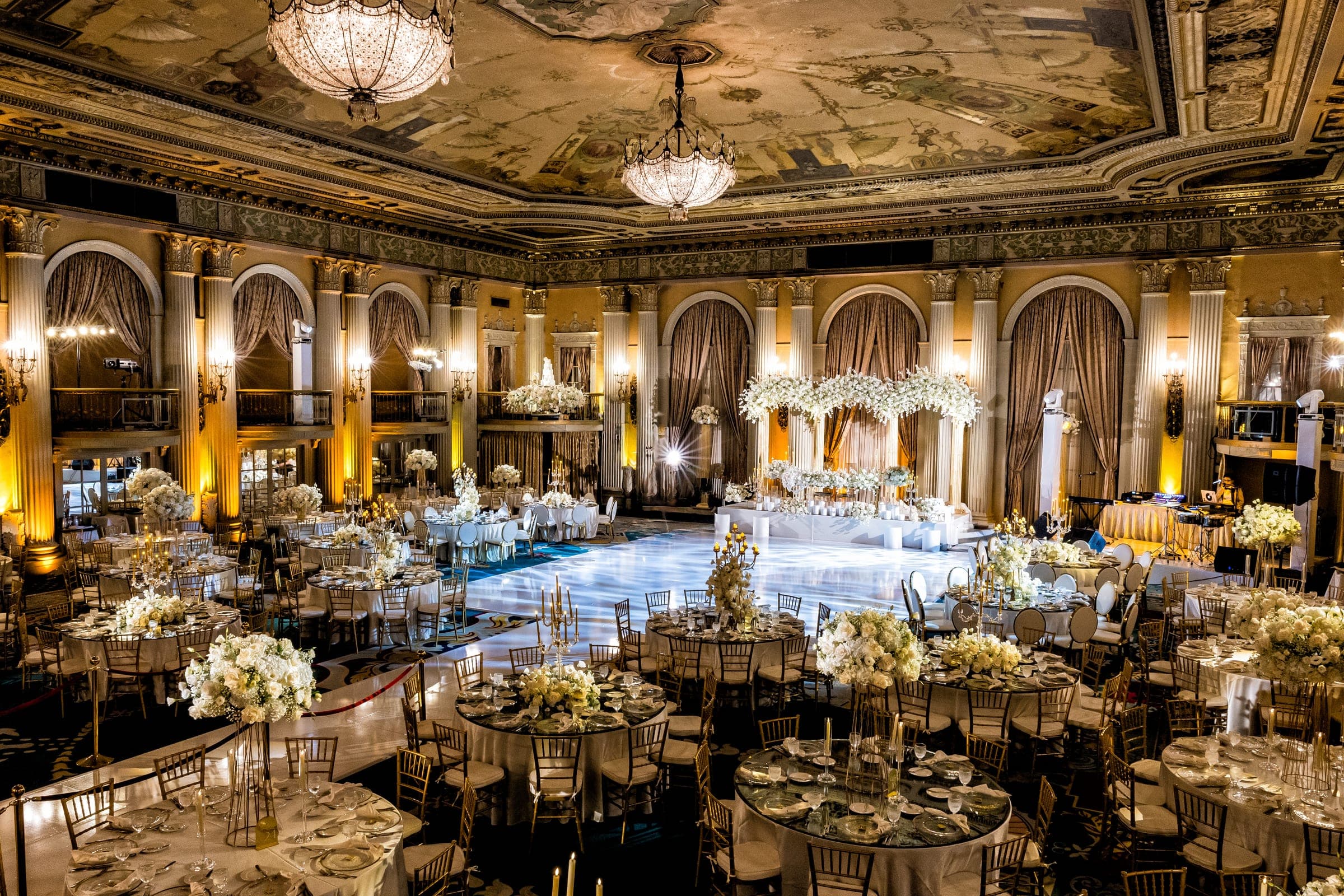 Grand ballroom set with round tables, gold chairs, tall floral centerpieces, chandeliers, and a floral stage backdrop.