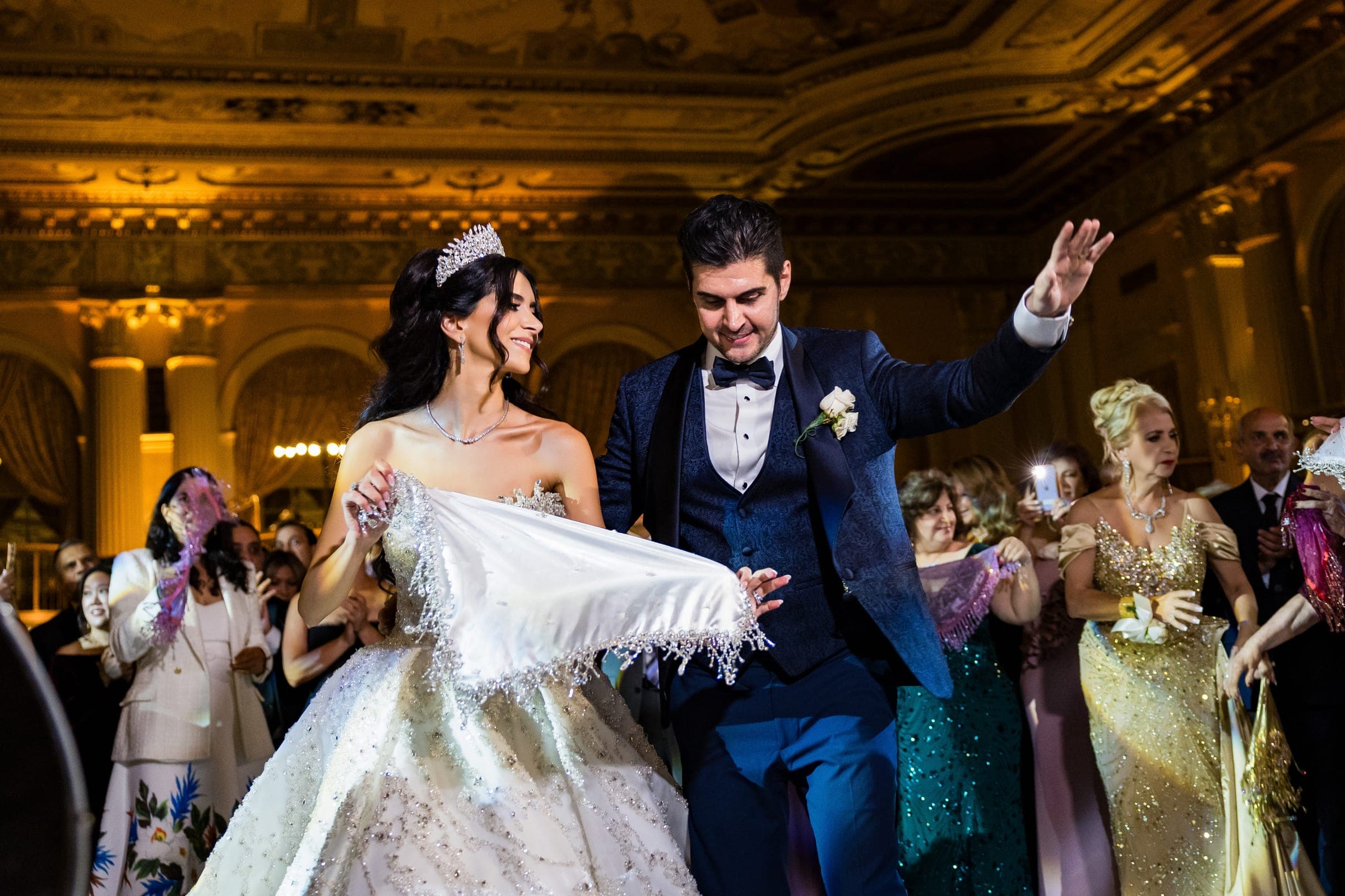 A bride in a tiara and groom in navy dance joyfully surrounded by cheering guests in an ornate ballroom.