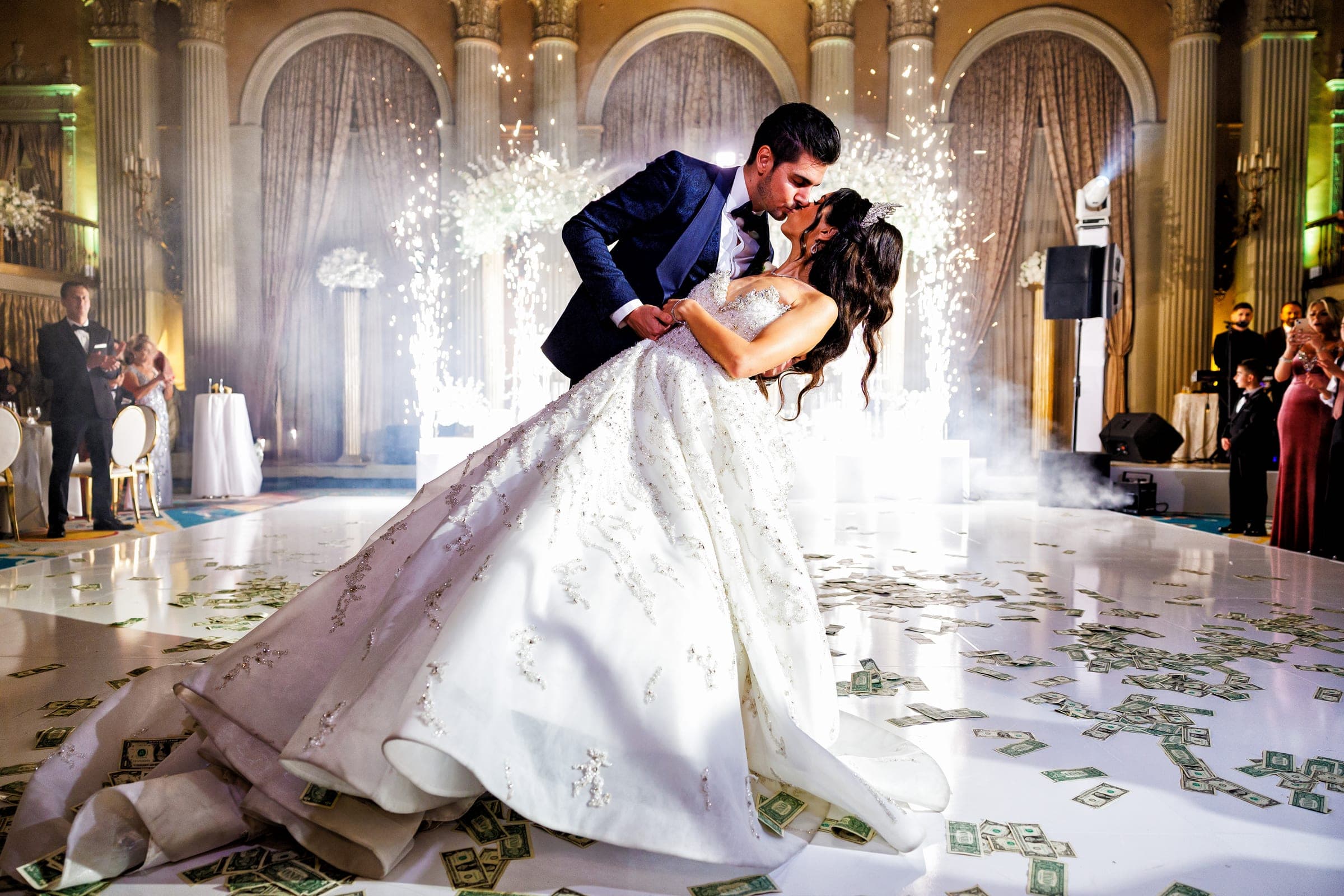 The groom dips and kisses his bride on a white dance floor scattered with money, sparklers glowing behind them.