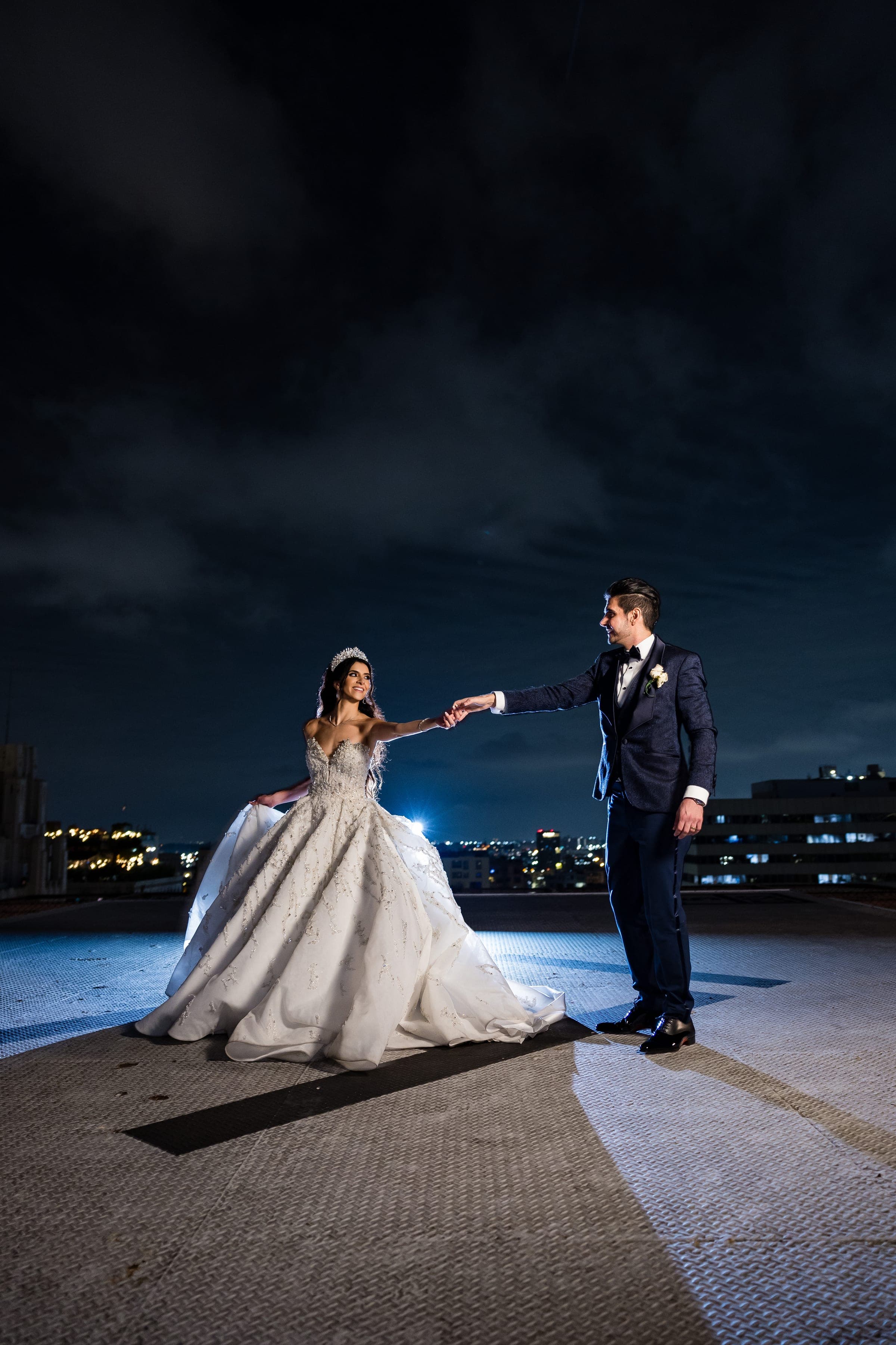 Bride and groom hold hands on a rooftop at night with the city skyline and dark clouds behind them.