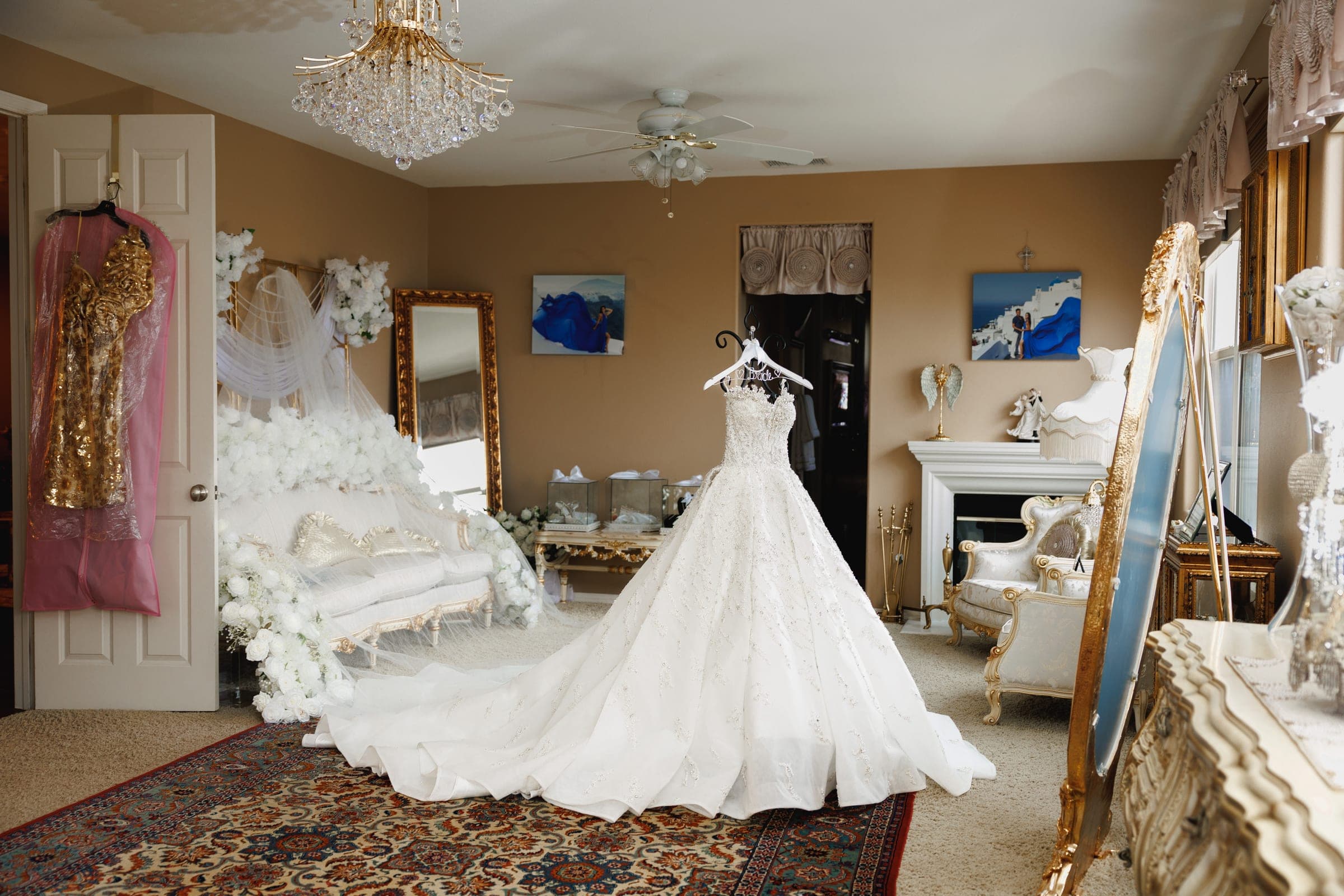 An ornate wedding gown hangs on display in a gilded room with chandelier, mirrors, and floral decor.